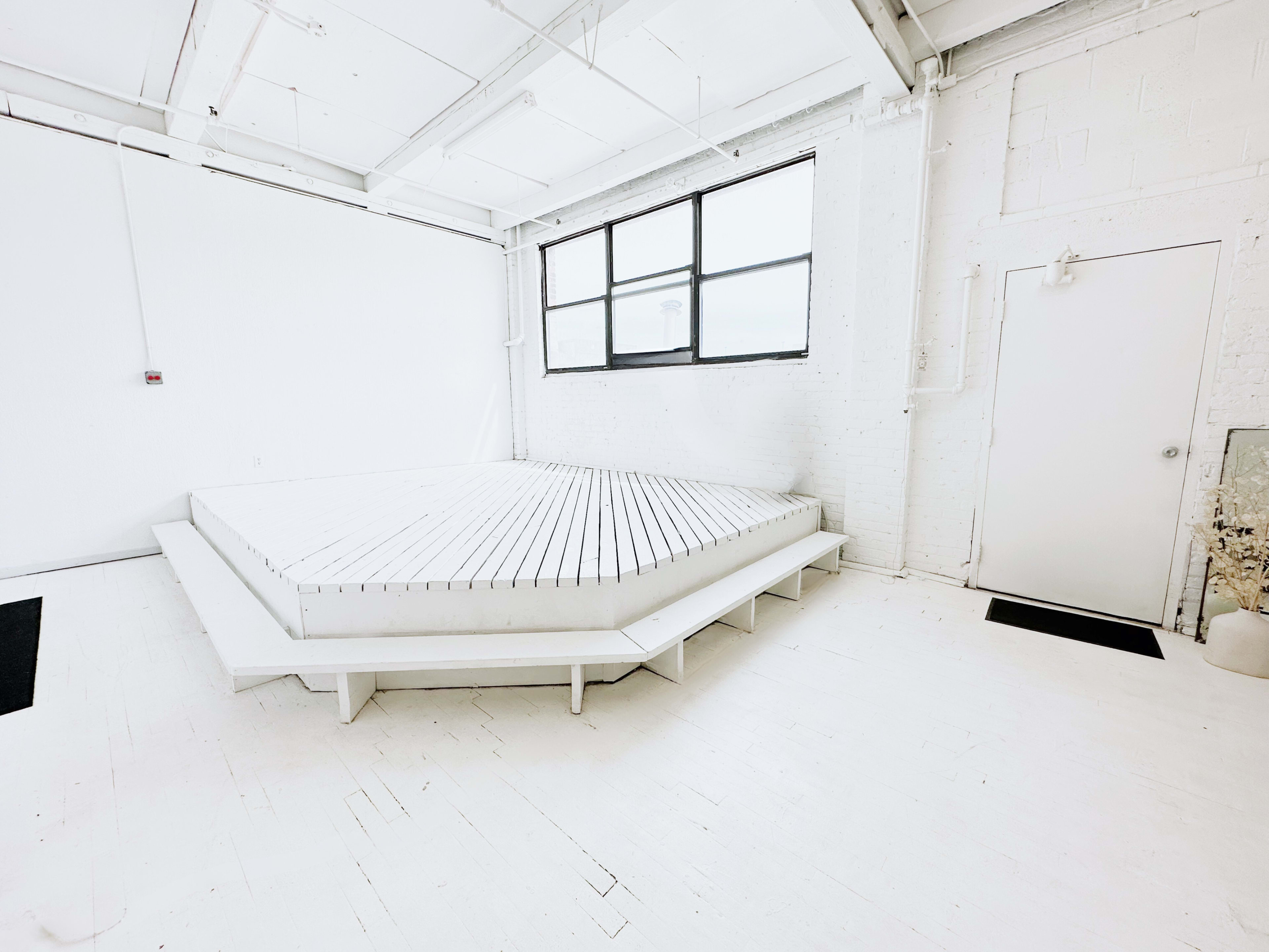 The image shows a minimalist, bright room featuring a large, white, elevated platform with slatted surfaces, situated near a window and a plain white door.