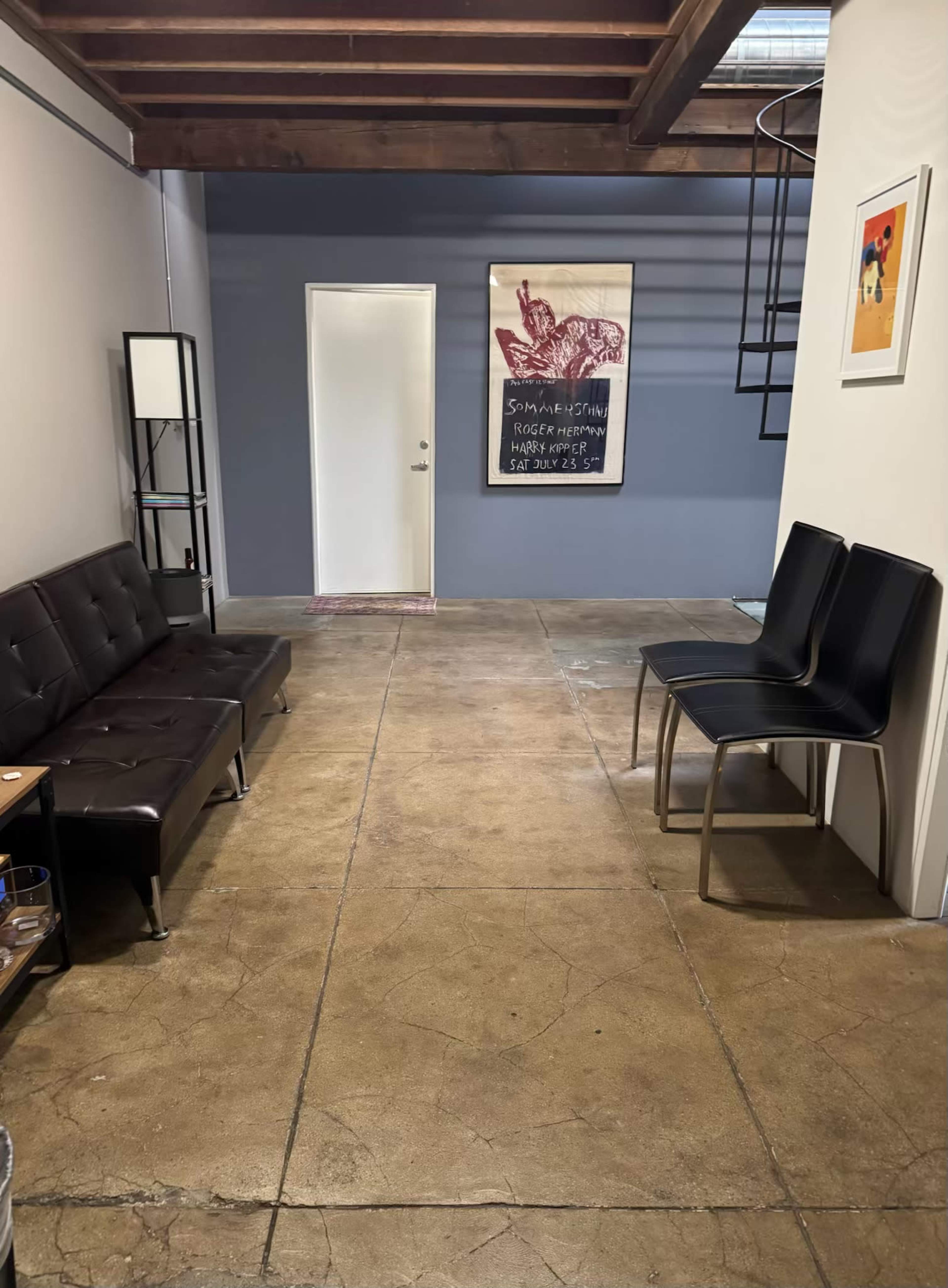 The image shows a narrow hallway with a dark sofa on the left, a few black chairs on the right, and a closed door at the end, along with an art piece on the wall.