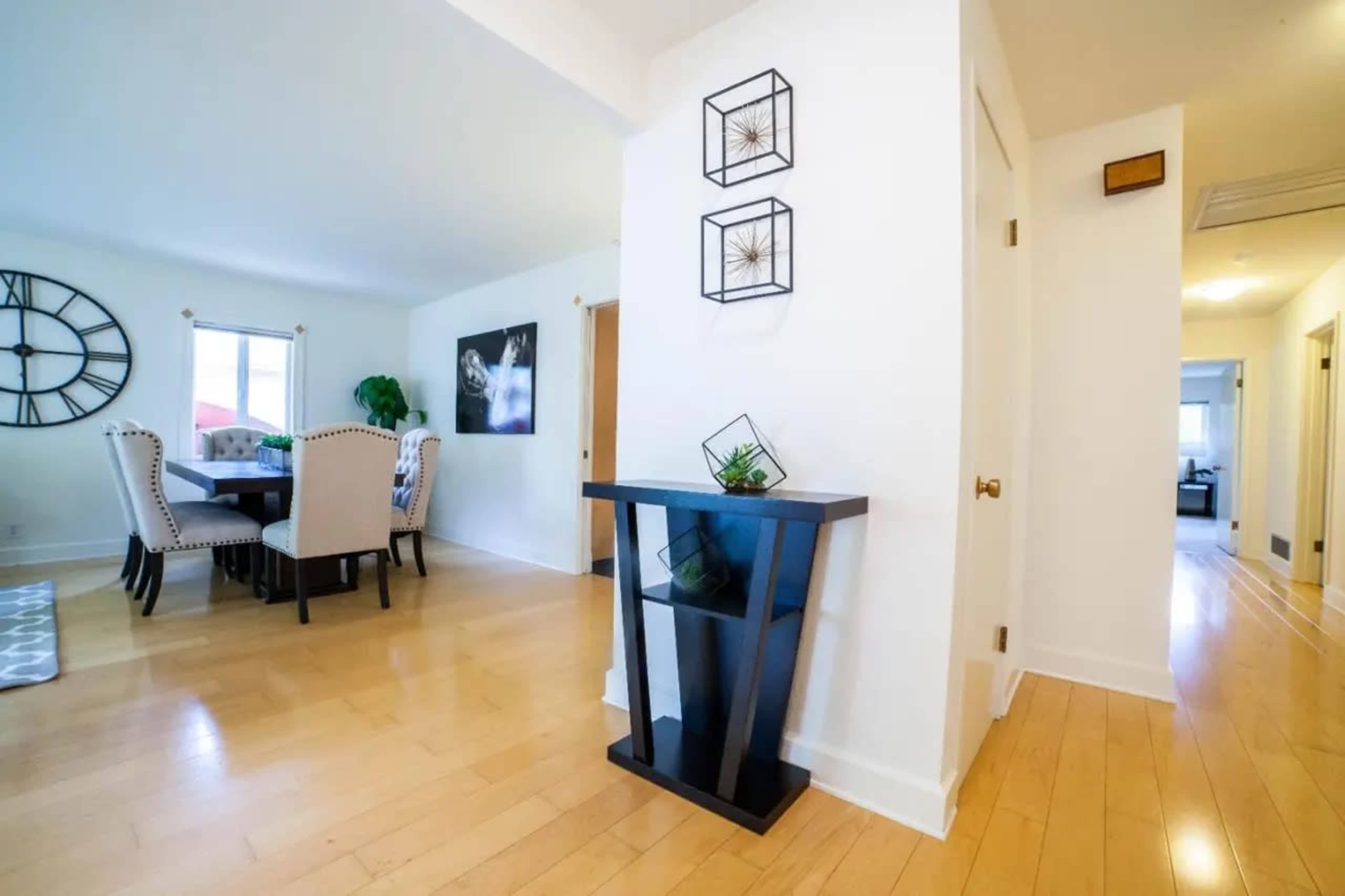 A hallway leading to a dining area with a table and chairs, featuring a black decorative stand with a plant and wall art in the background.