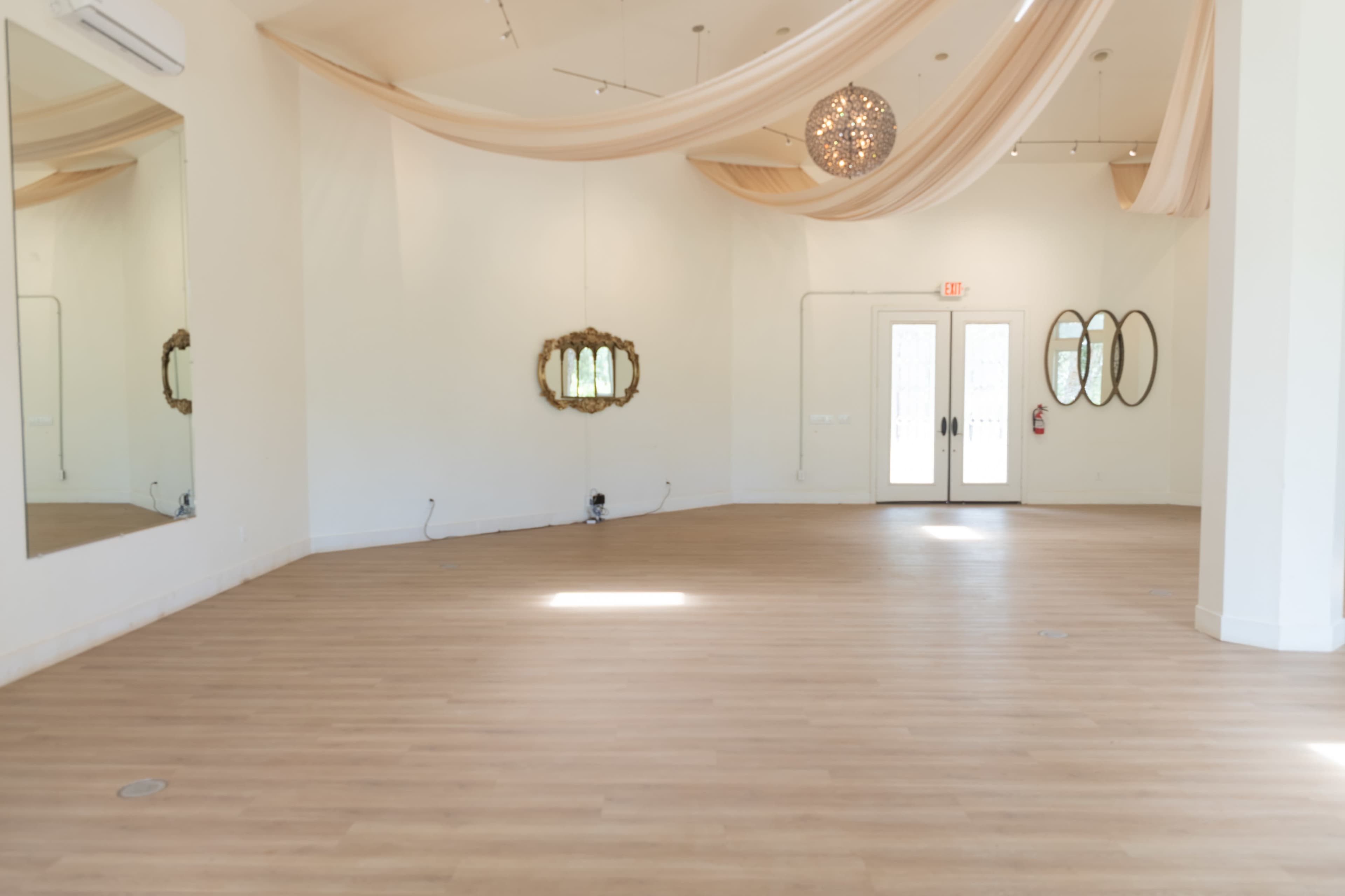 The image shows a spacious interior room with light wooden flooring, a pair of glass doors, decorative mirrors on the walls, and fabric draping from the ceiling.