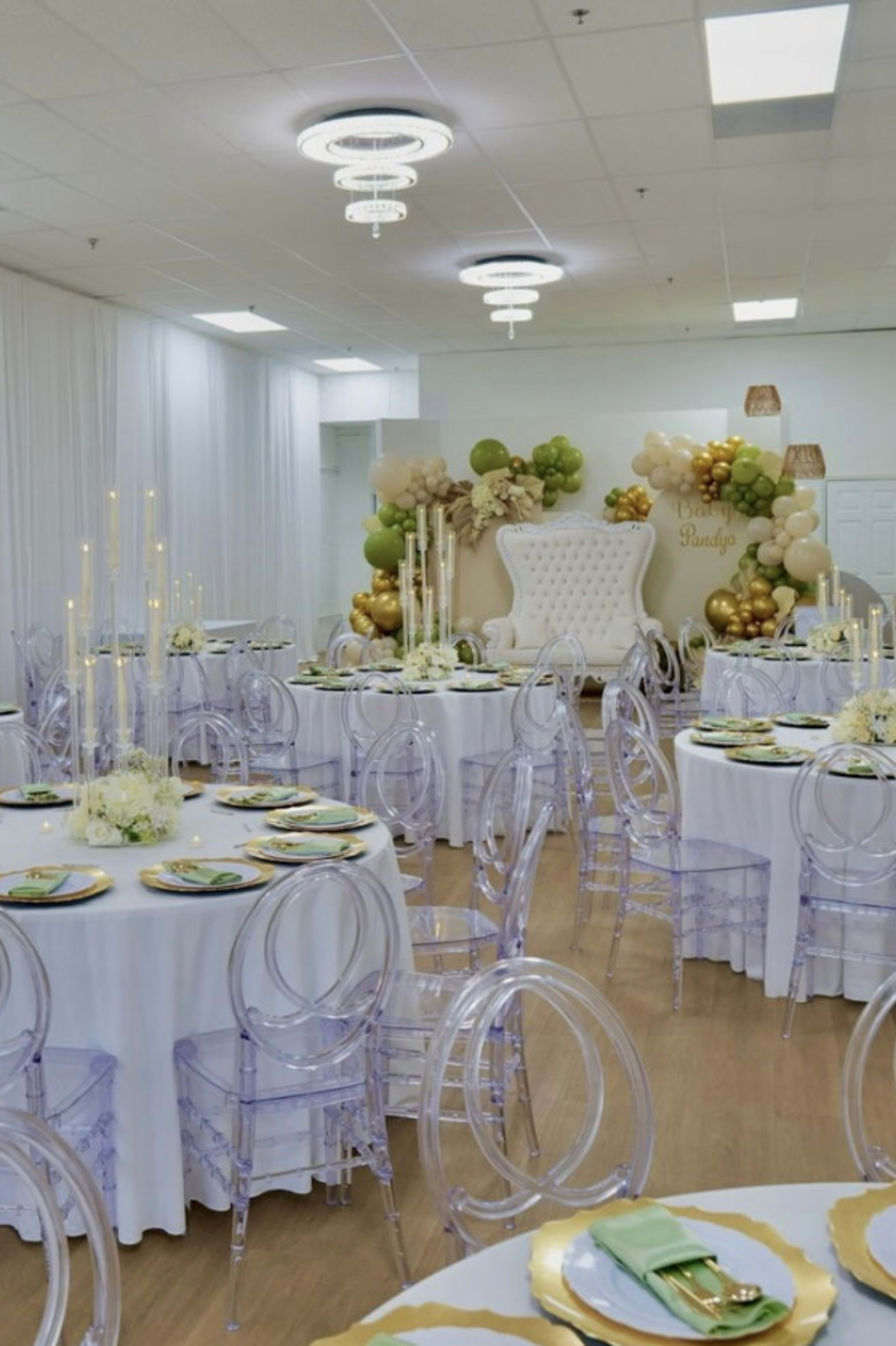 The image shows a banquet hall decorated with round tables featuring transparent chairs, white tablecloths, and gold and green balloon arrangements in the background.