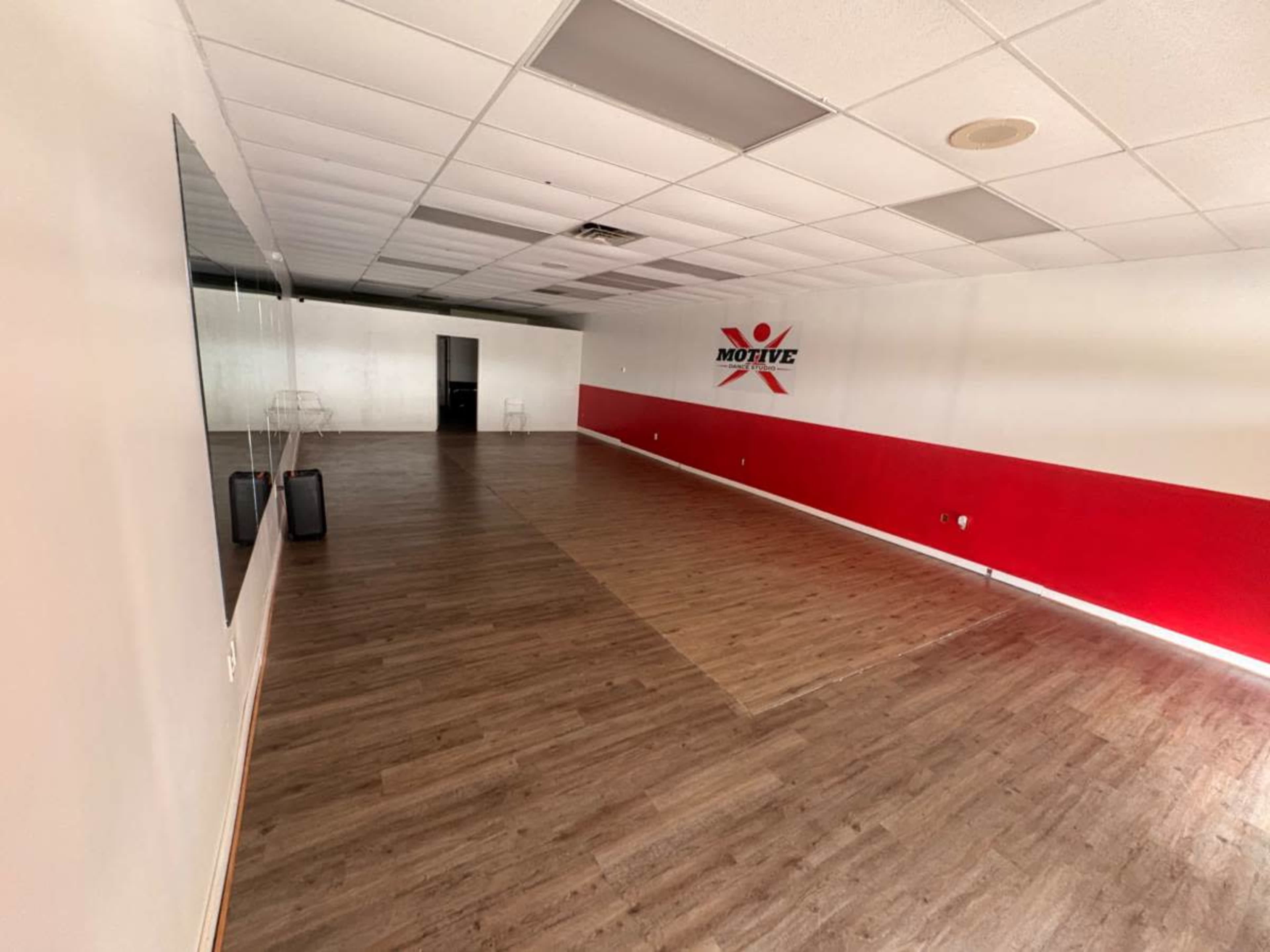 The image shows an empty interior space with wooden flooring, a red accent wall, and mirrors on one side.