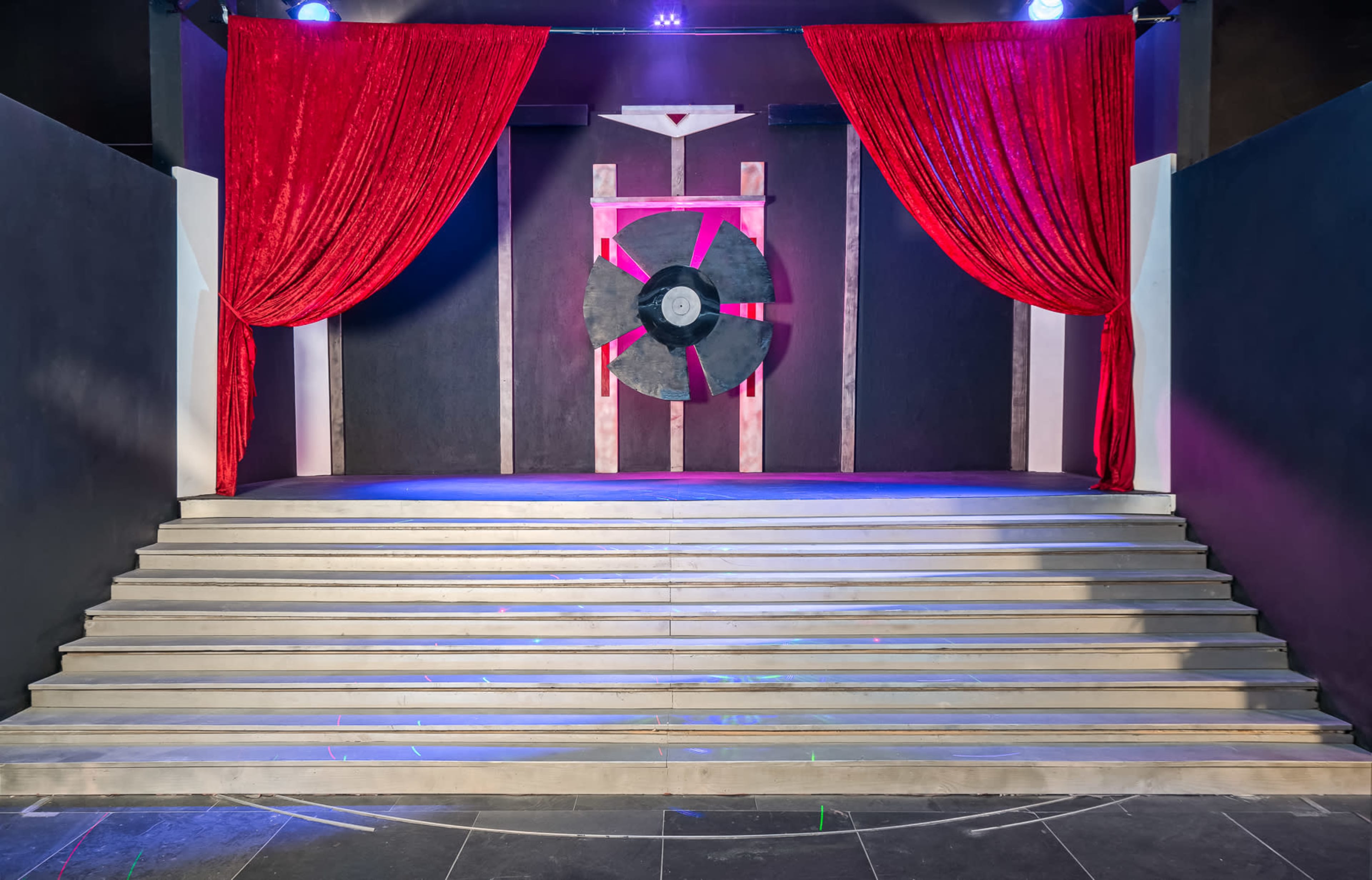 A stage with red curtains and a large fan backdrop is set against a dark wall, featuring stepped flooring leading up to it.