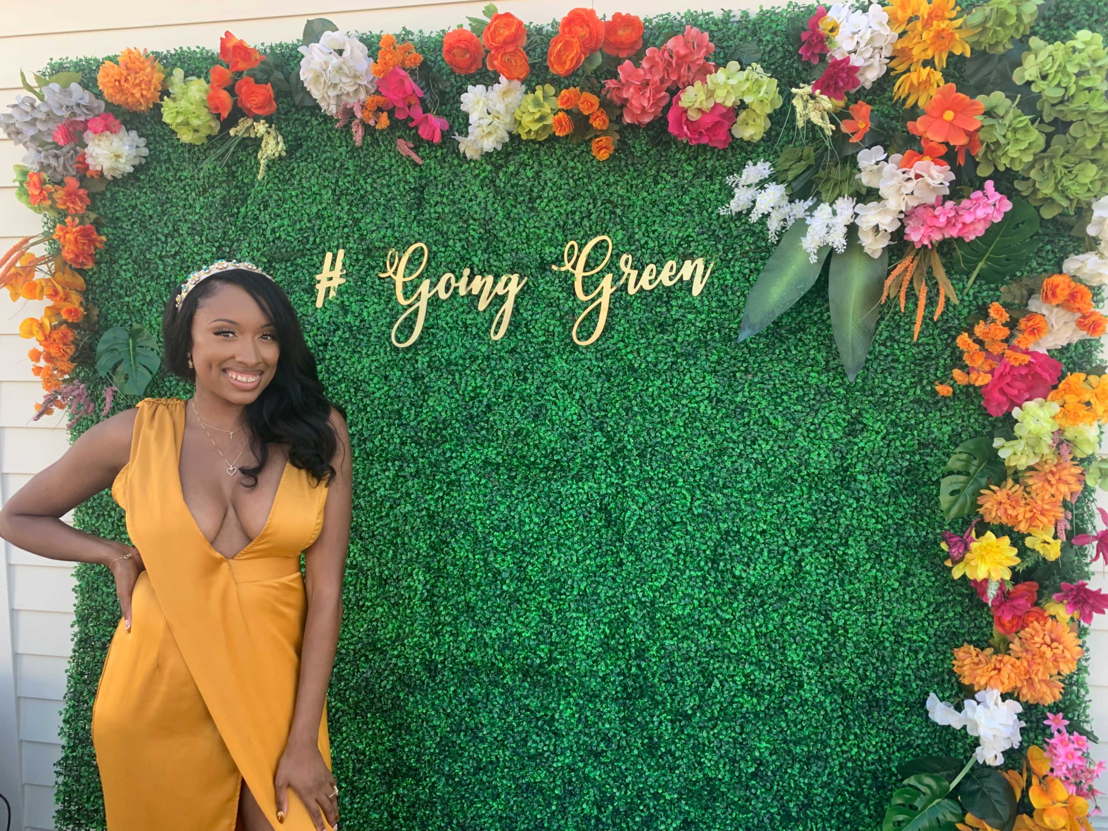 A woman in a yellow dress poses next to a green floral backdrop with the text "# Going Green" displayed in gold letters.