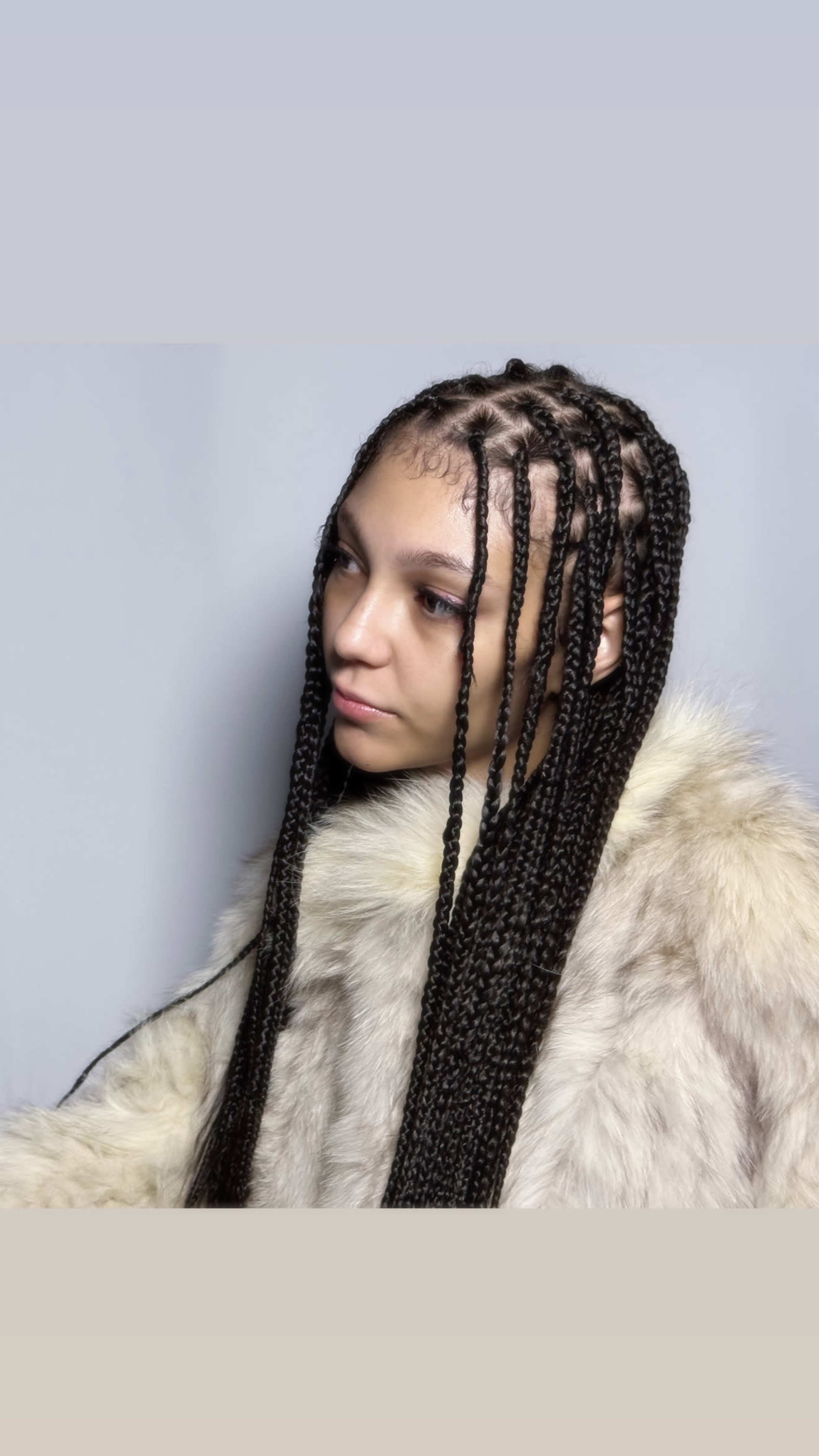 A person with long braided hair is seen wearing a fur coat against a light gray background.