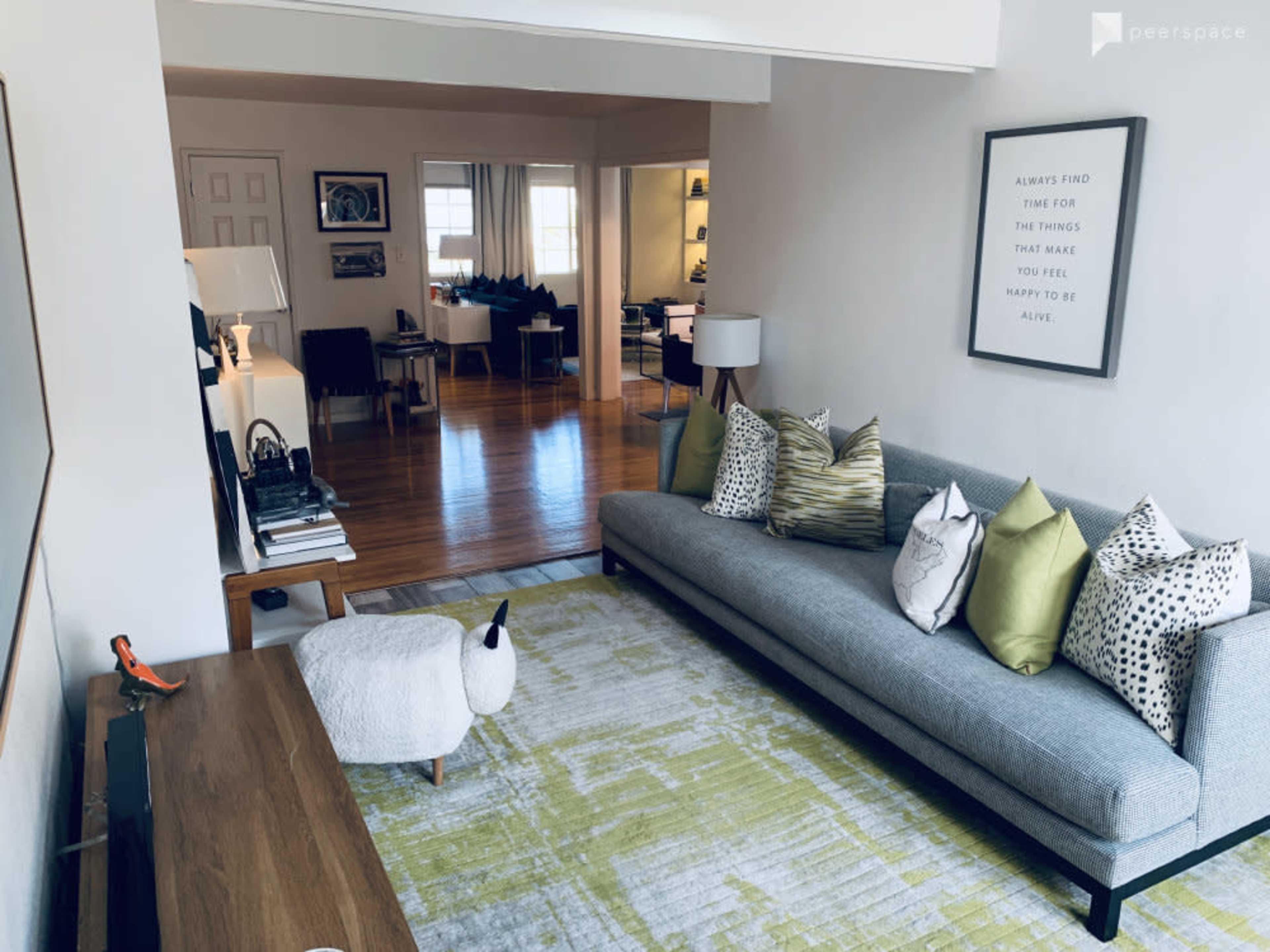 A living room features a gray sofa adorned with decorative pillows, a patterned rug, and a minimalist artwork on the wall, with a dining area visible in the background.
