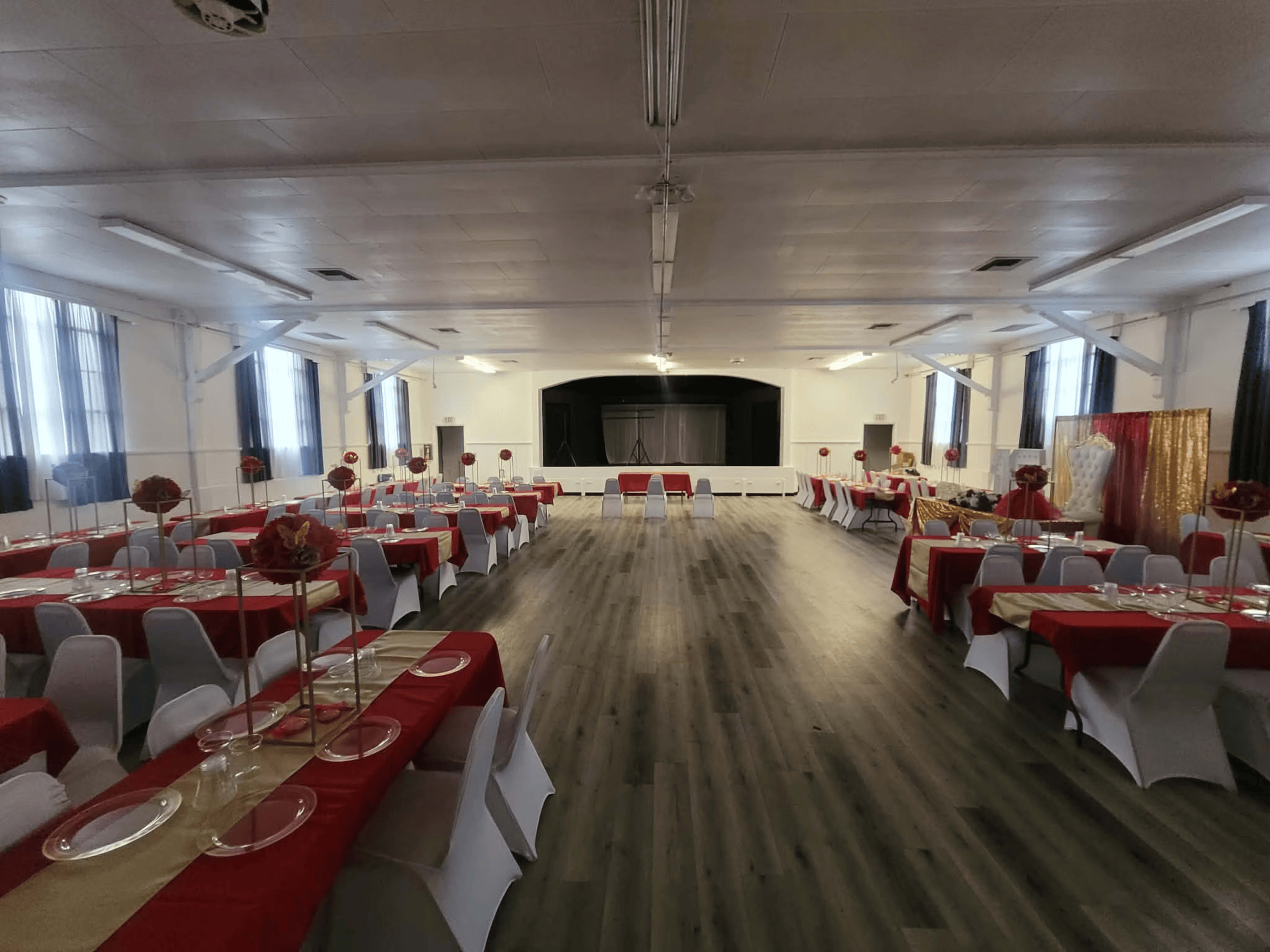 The image shows a spacious indoor event hall set up with red and white table arrangements, featuring round tables adorned with centerpieces, and a stage at the front.