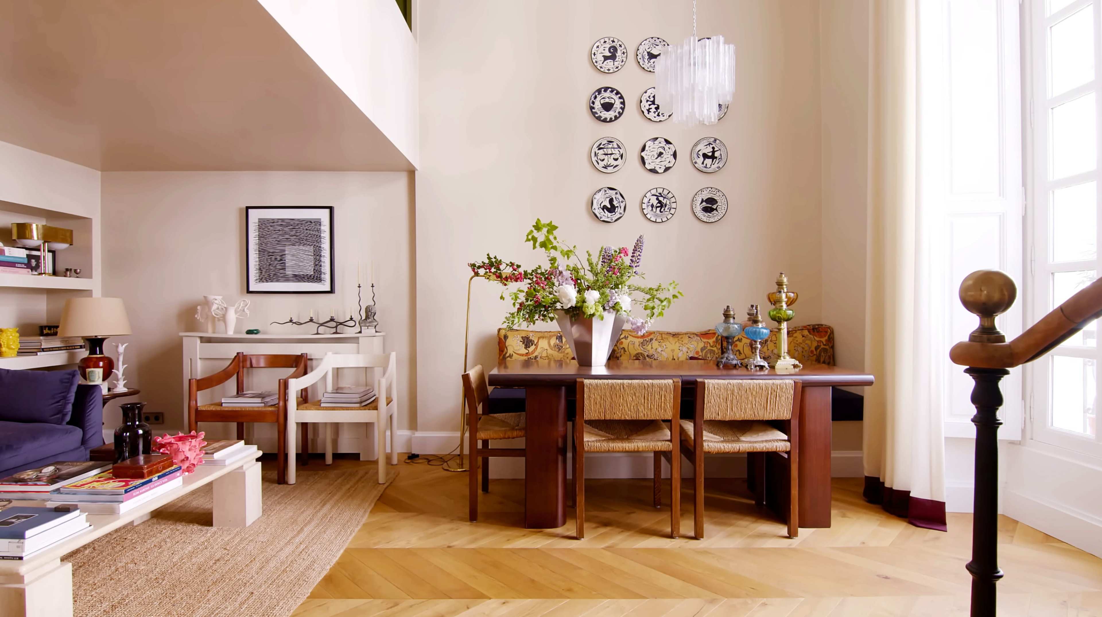 A dining area features a wooden table surrounded by chairs, adorned with a vase of flowers and decorative plates on the wall.
