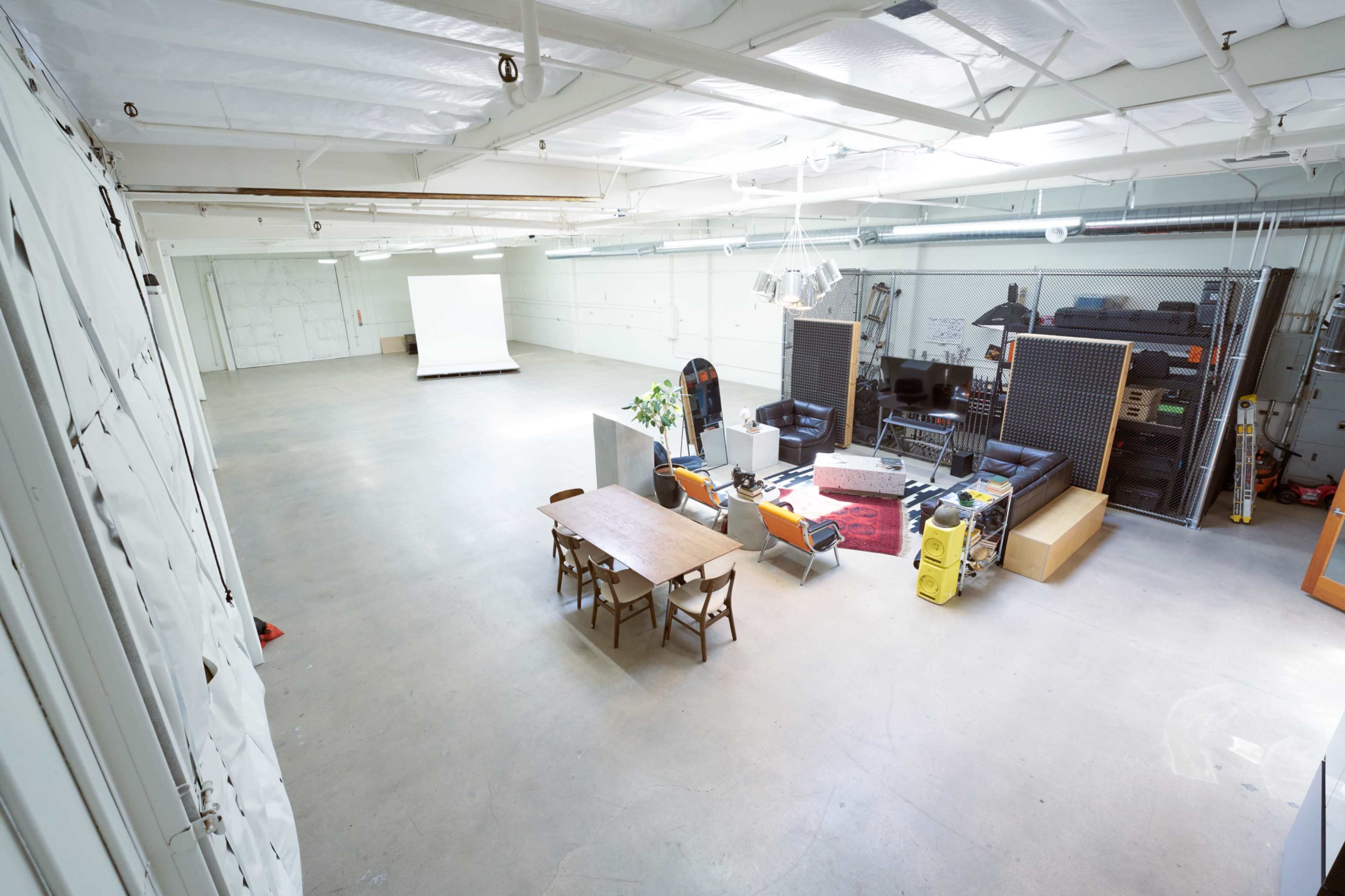 The image shows a large, empty warehouse-style space with a table and chairs, a projection screen, and various storage items scattered around.