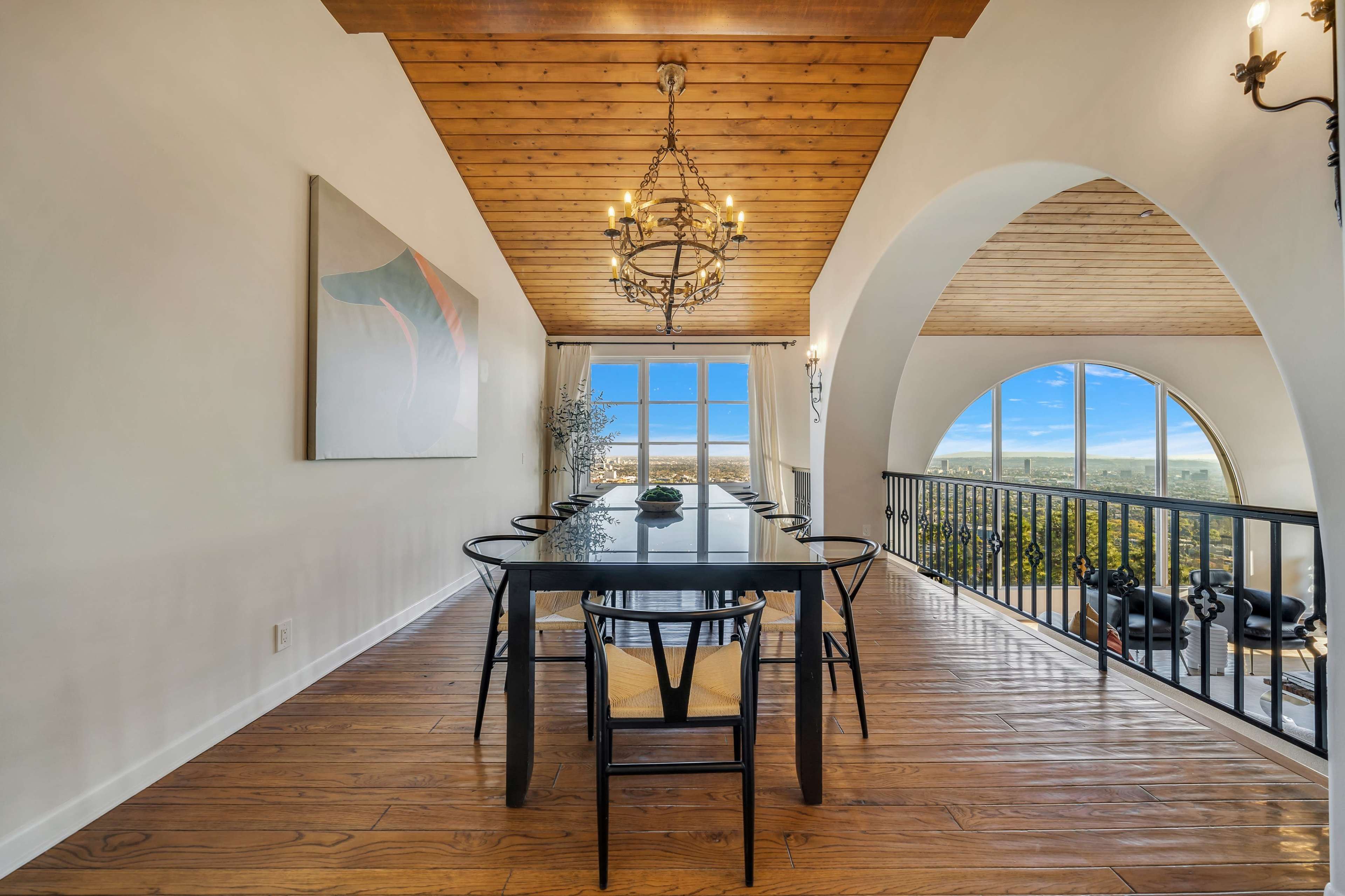 The image shows a dining area with a long black table surrounded by chairs, large windows offering a view, and a wooden ceiling with a chandelier above.