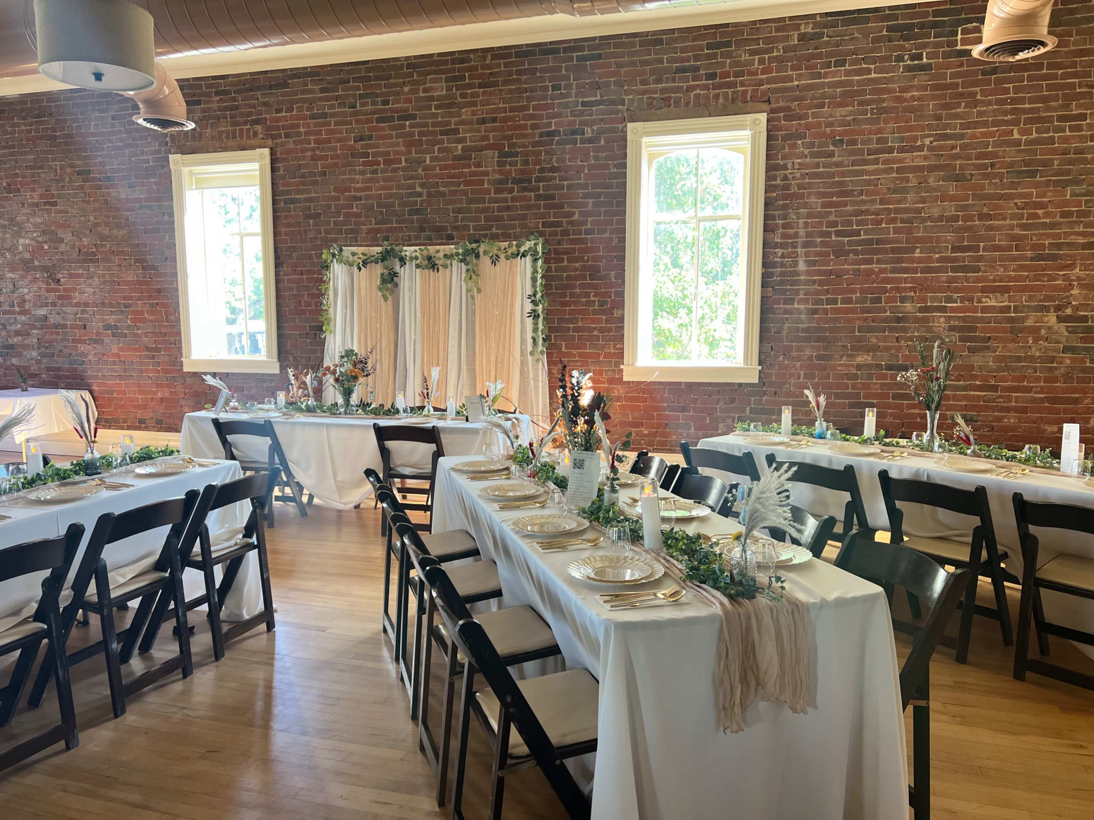 The image shows a decorated event space featuring long tables set for a gathering, with a backdrop of greenery and white drapery against a brick wall.