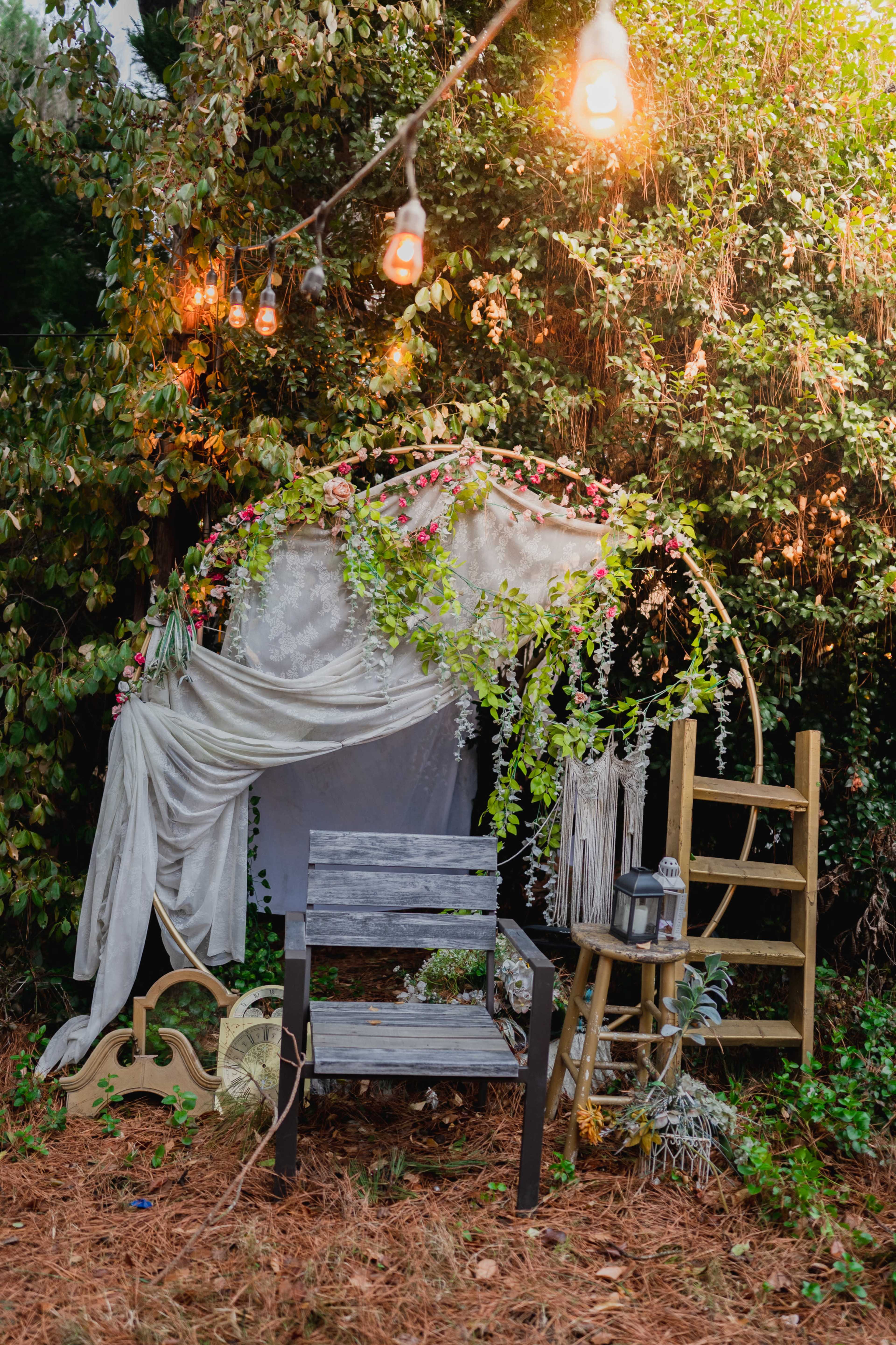 A rustic seating area adorned with floral decorations, a draped fabric canopy, and a wooden ladder, set against a backdrop of greenery.