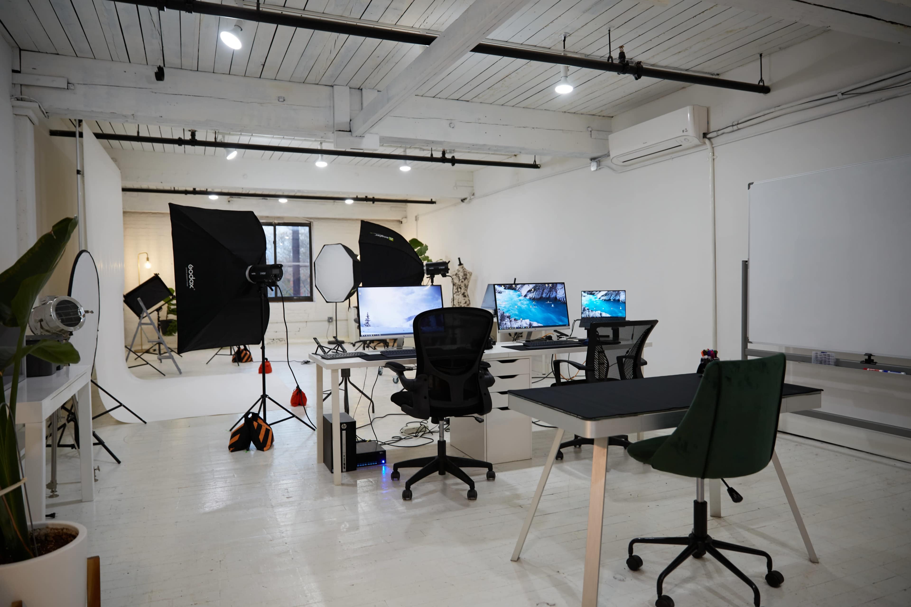 A modern workspace with multiple monitors, office chairs, and studio lighting equipment in a well-lit room with white walls and wooden beams.
