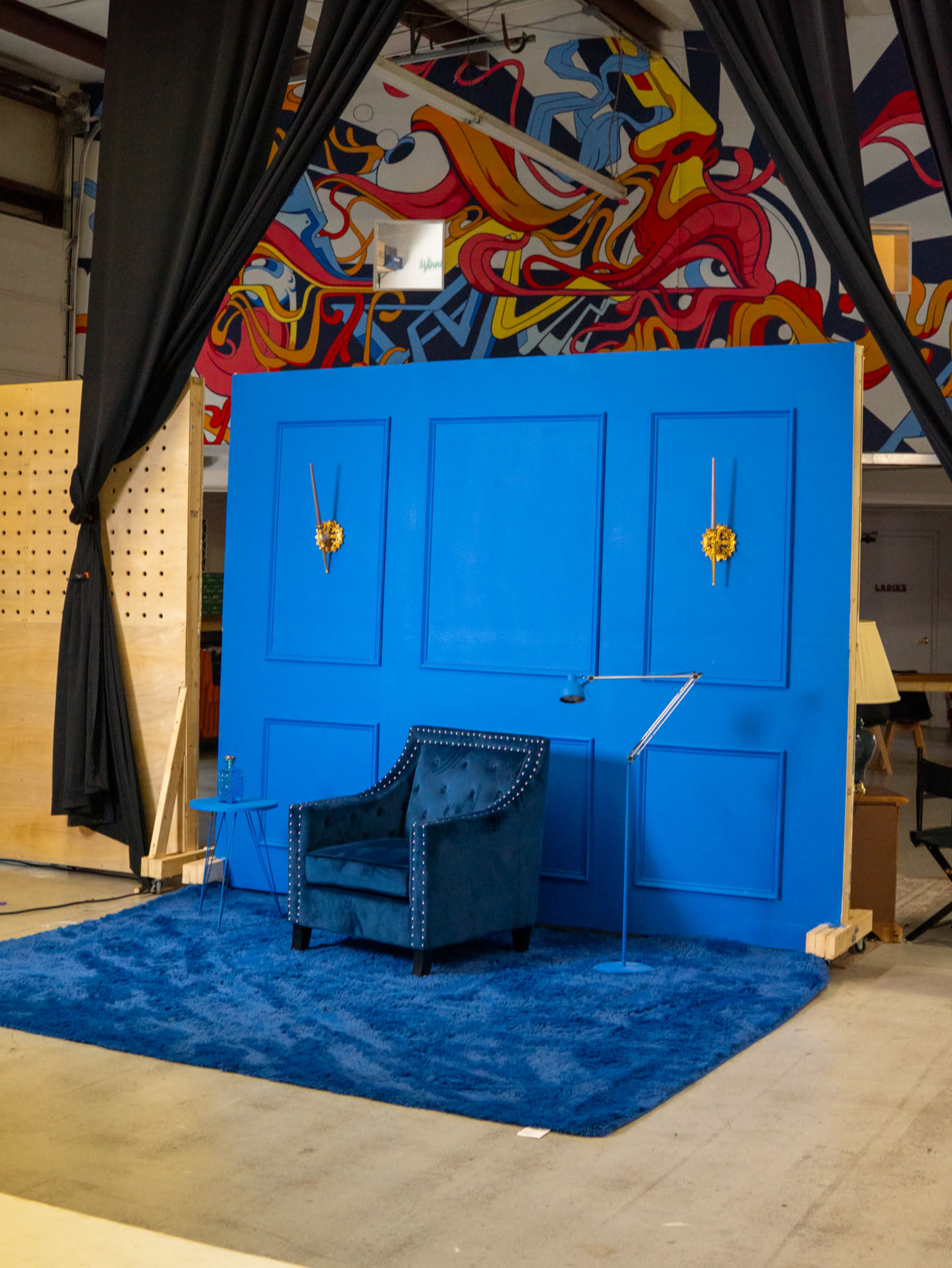 A bright blue wall serves as a backdrop for a velvet chair and a small side table, set on a blue rug, in a spacious indoor area featuring abstract wall art.