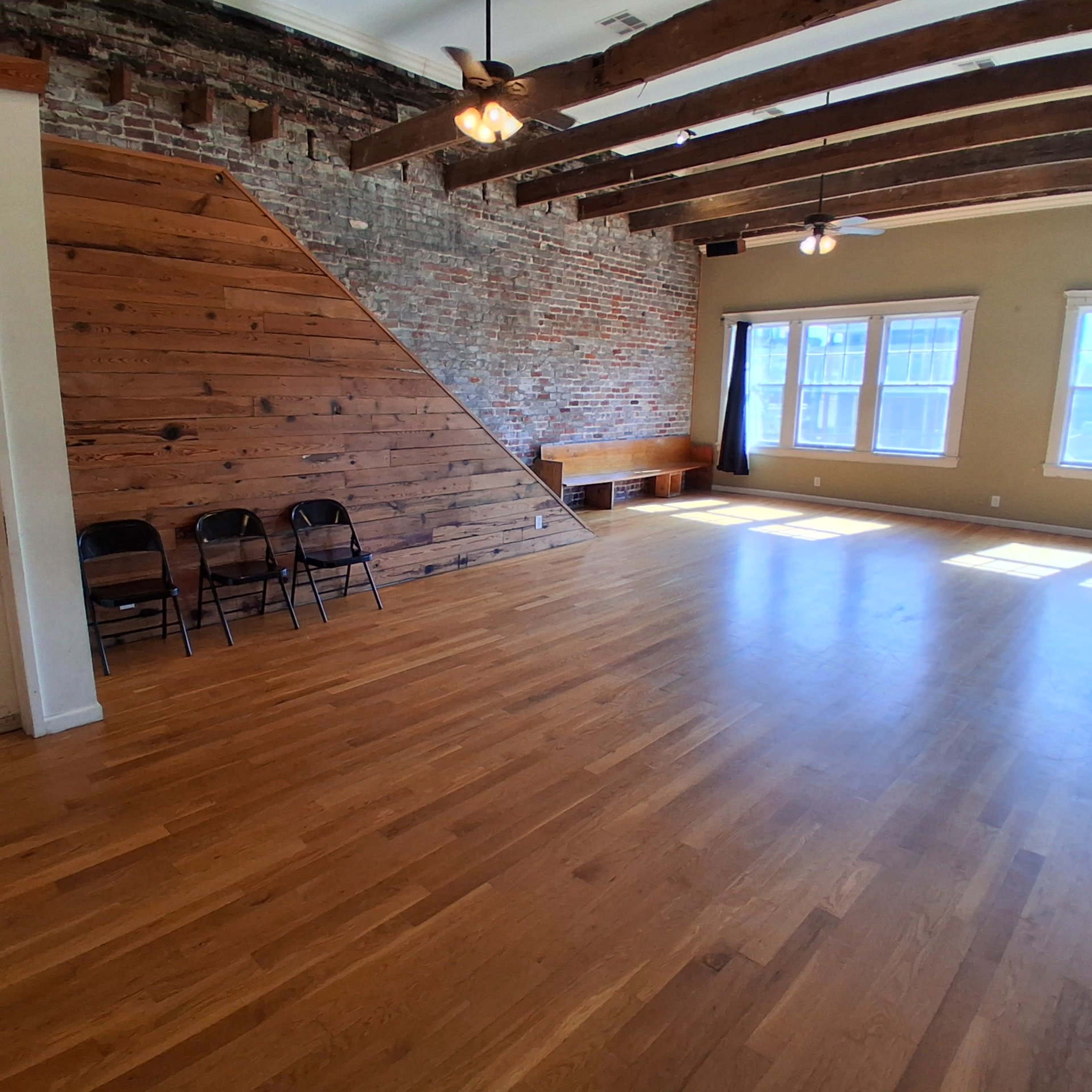 The image shows a spacious room with wooden floors, exposed brick walls, wooden beams, and a few folding chairs against one wall.