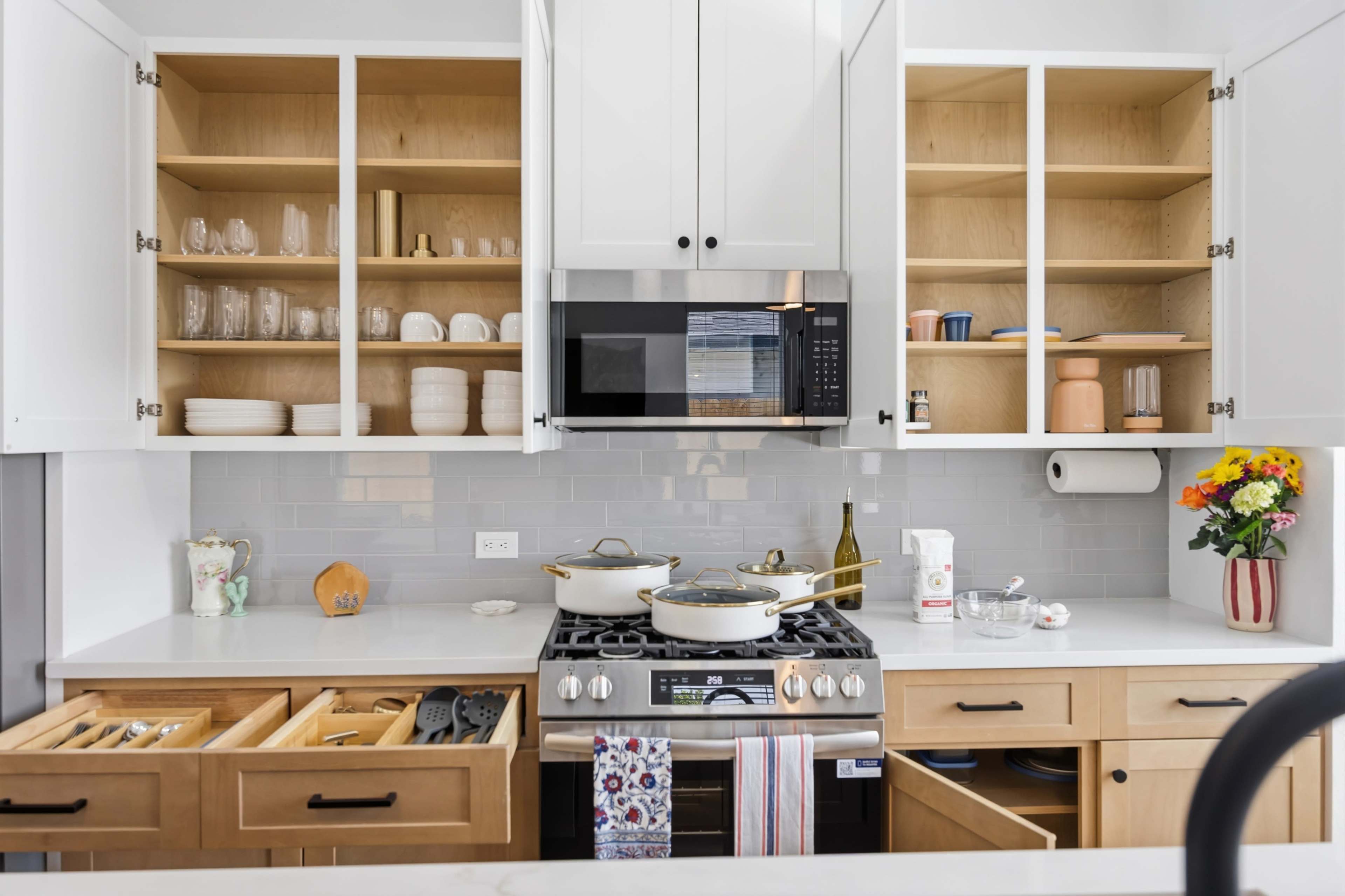 A modern kitchen features white cabinetry, open shelves displaying glassware and dishware, a stainless steel microwave, and a gas stove with pots and a decorative flower arrangement.