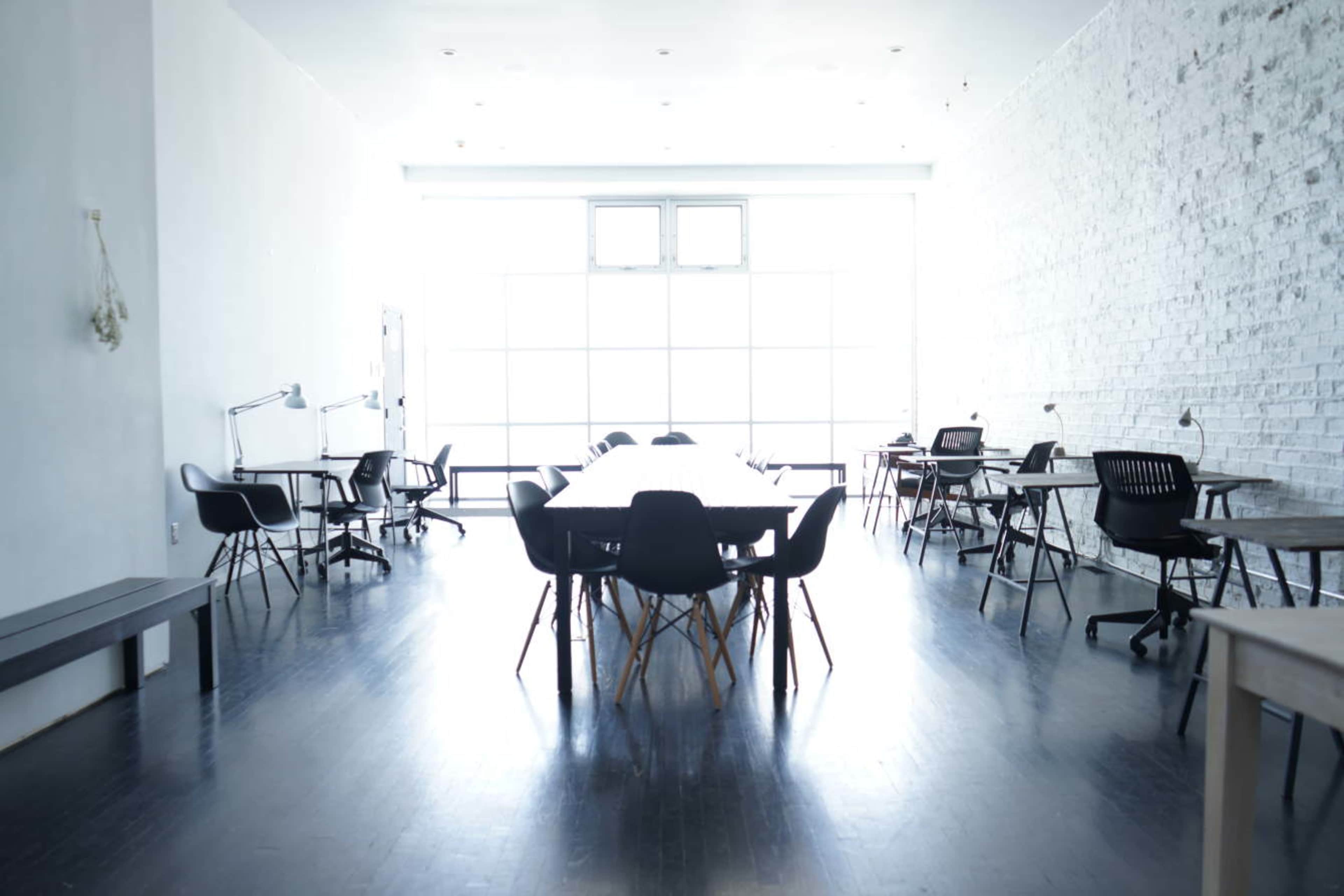 A spacious, well-lit room with a long table surrounded by black chairs, and several workstations along the walls.