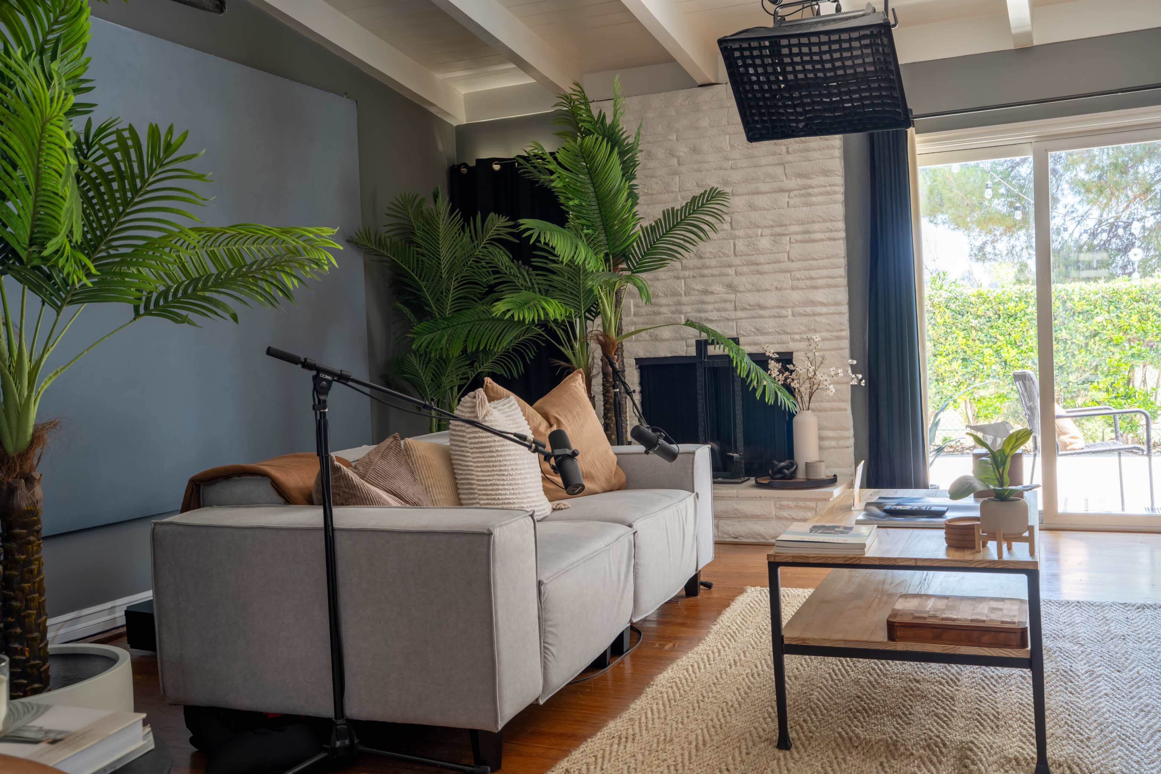 A modern living room features a grey sofa with pillows, tropical plants, a wooden coffee table, and a stone fireplace, with sliding glass doors leading to an outdoor area.