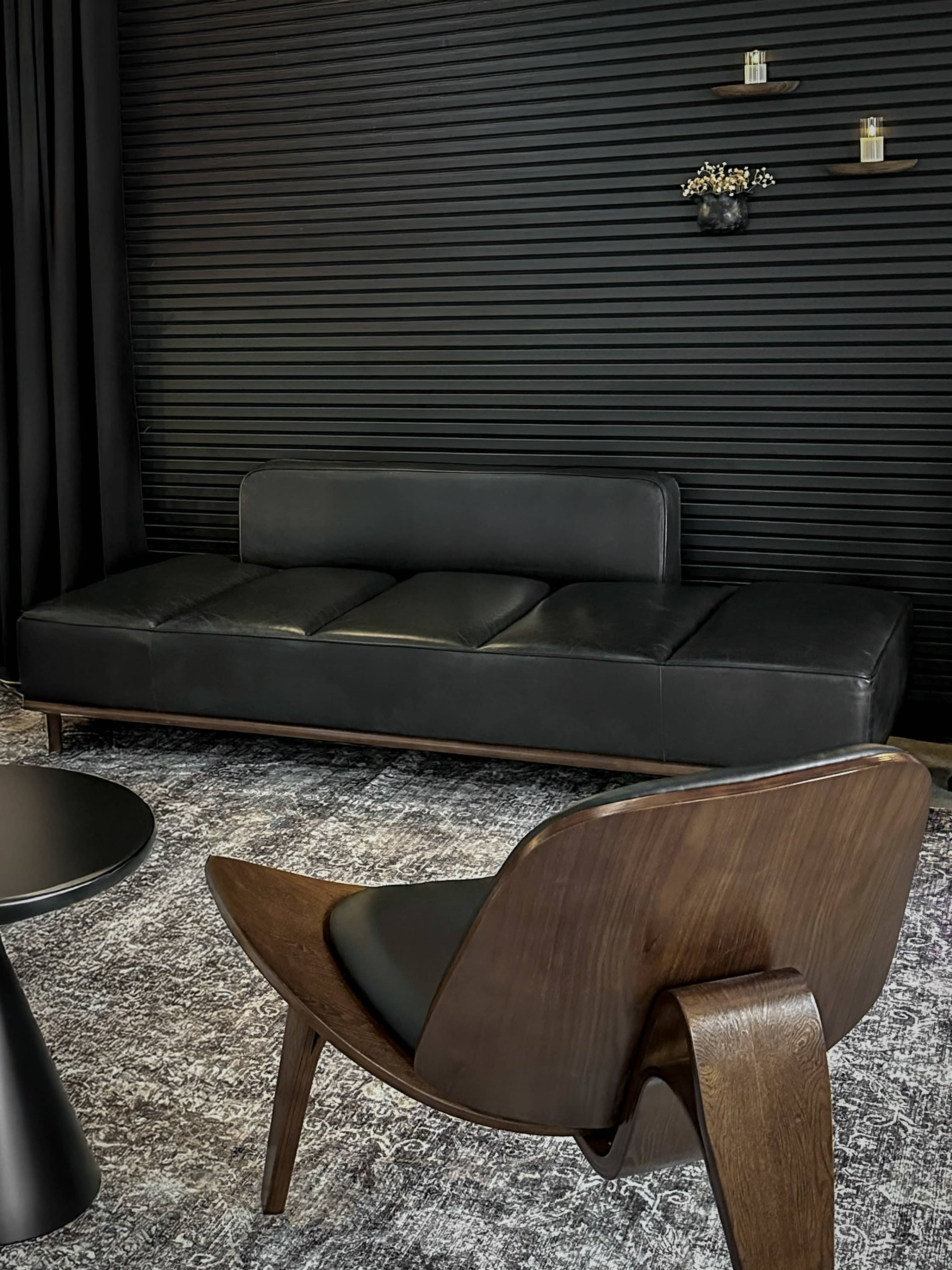 A modern black leather sofa is positioned in front of a wooden chair, against a dark striped wall with decorative shelves holding small items.