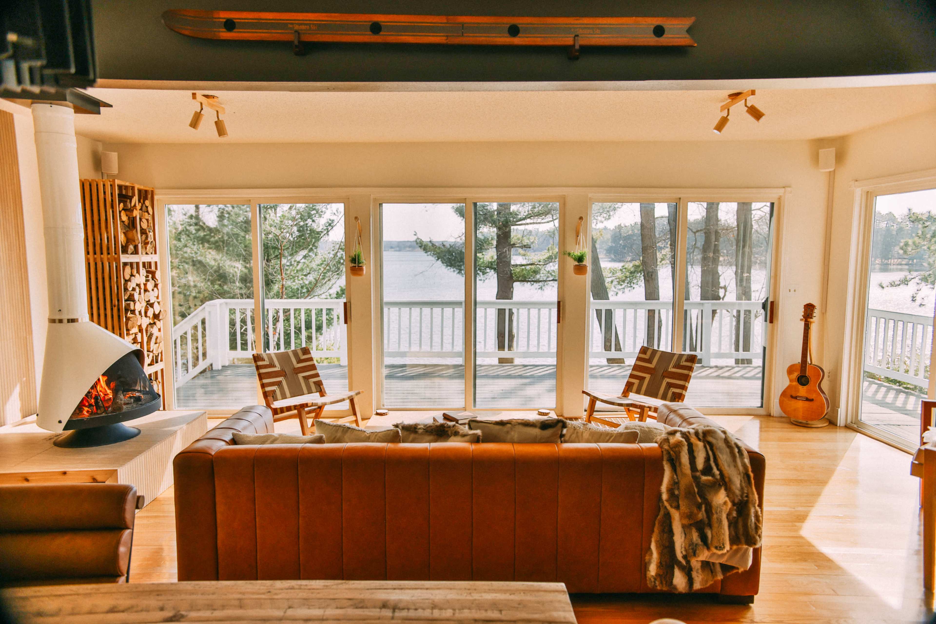 A modern living room features large windows overlooking water, with a leather sofa, wooden chairs, and a guitar positioned nearby.