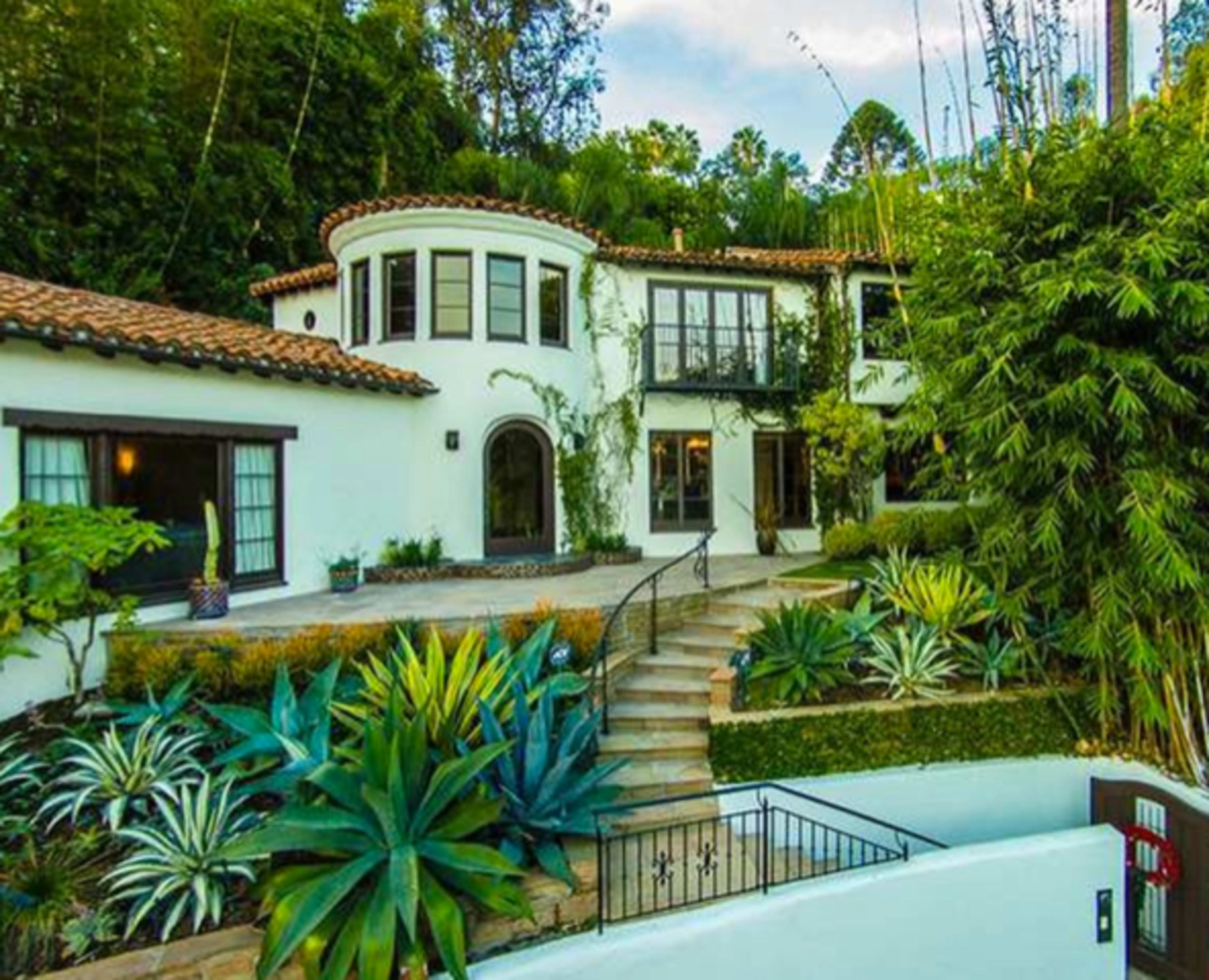 The image shows a two-story Mediterranean-style house with a curved balcony, surrounded by lush greenery and landscaping, including various succulents and steps leading up to the entrance.