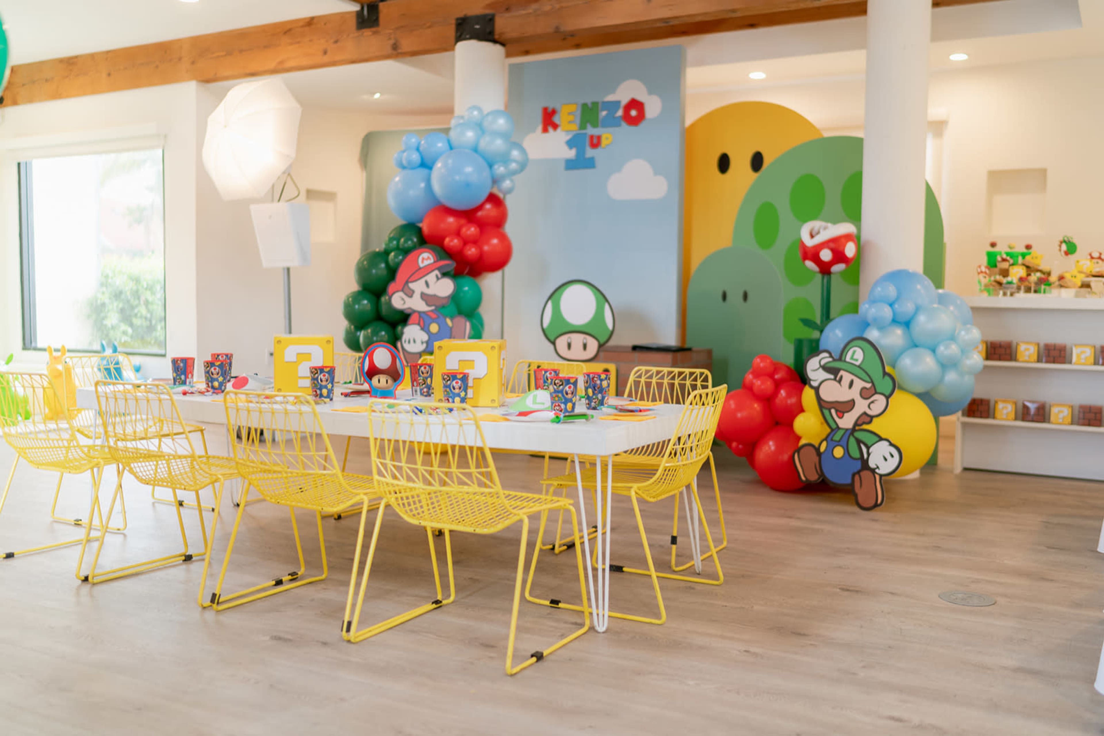 A colorful children's party setup featuring a table surrounded by yellow wire chairs, decorated with balloons and character cutouts from a video game theme.