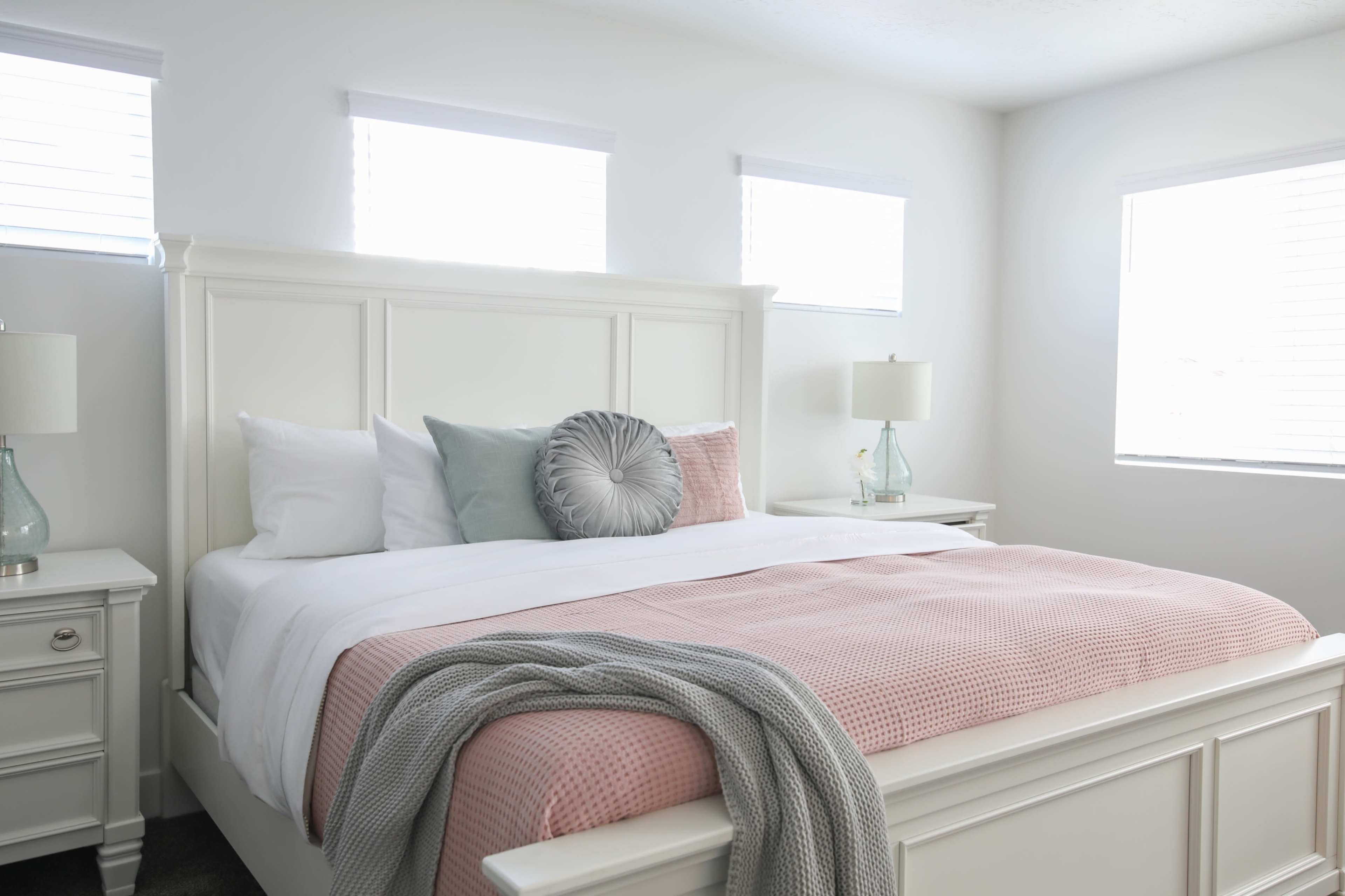 A neatly made bed with a pink bedspread and decorative pillows in a bright bedroom featuring two windows and matching bedside lamps.