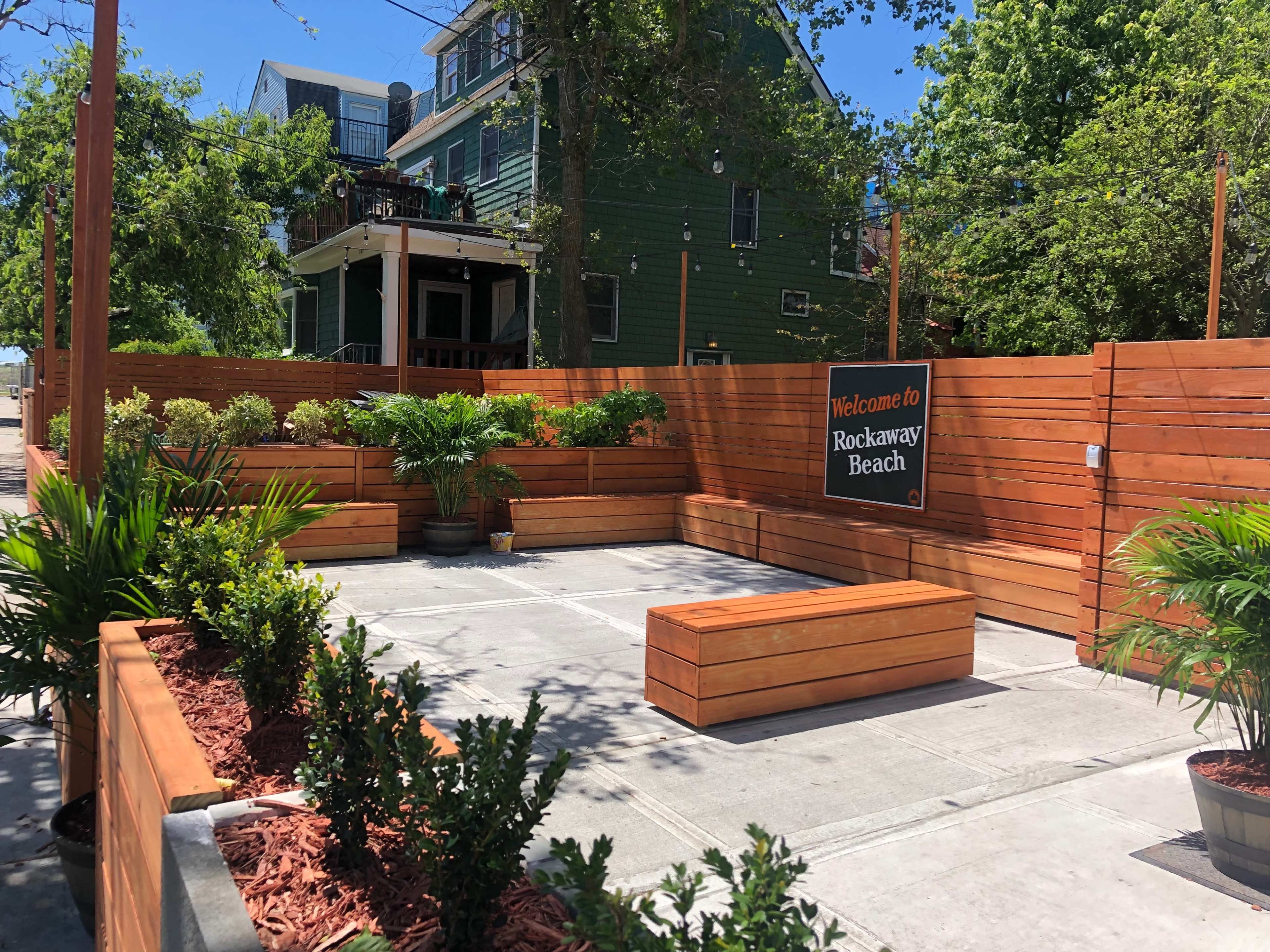 The image shows a landscaped outdoor space with wooden benches, potted plants, and a sign that reads "Welcome to Rockaway Beach."