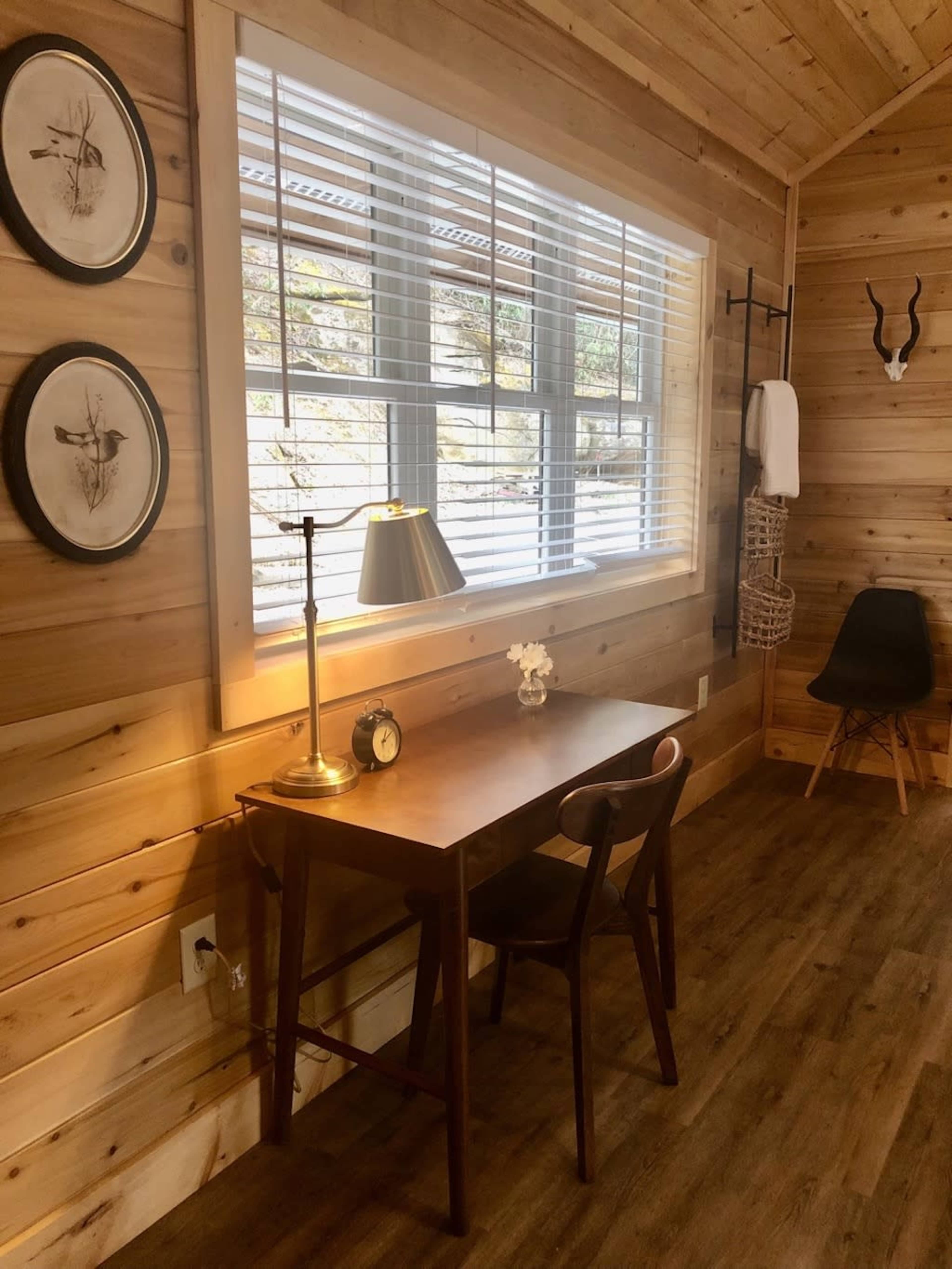 A minimalist workspace featuring a wooden desk with a lamp, a clock, and floral decor, alongside a chair and wall-mounted storage in a room with wooden paneling and a large window.