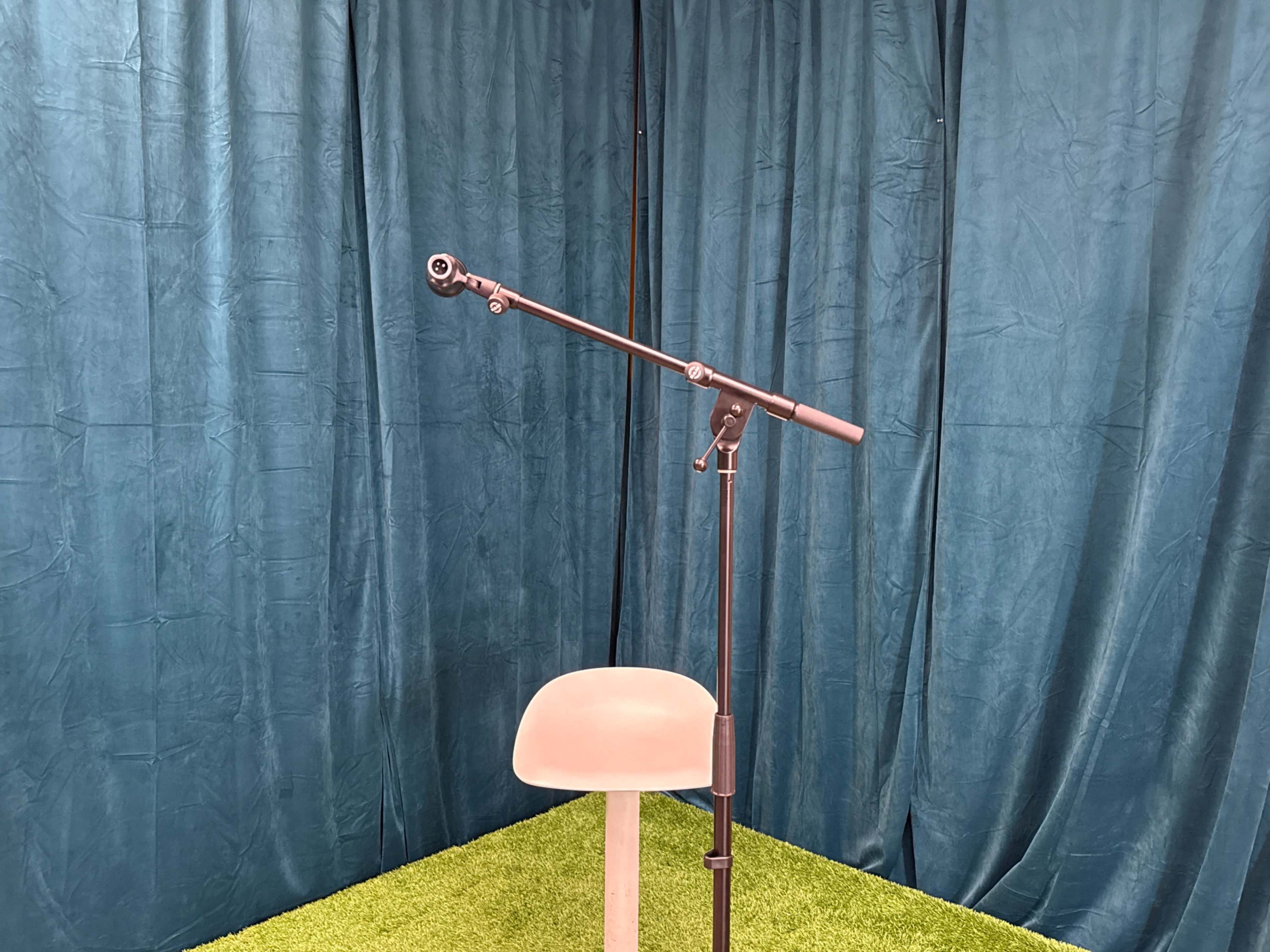 A microphone stand is positioned next to a simple chair on a grassy green surface, with a blue curtain backdrop.