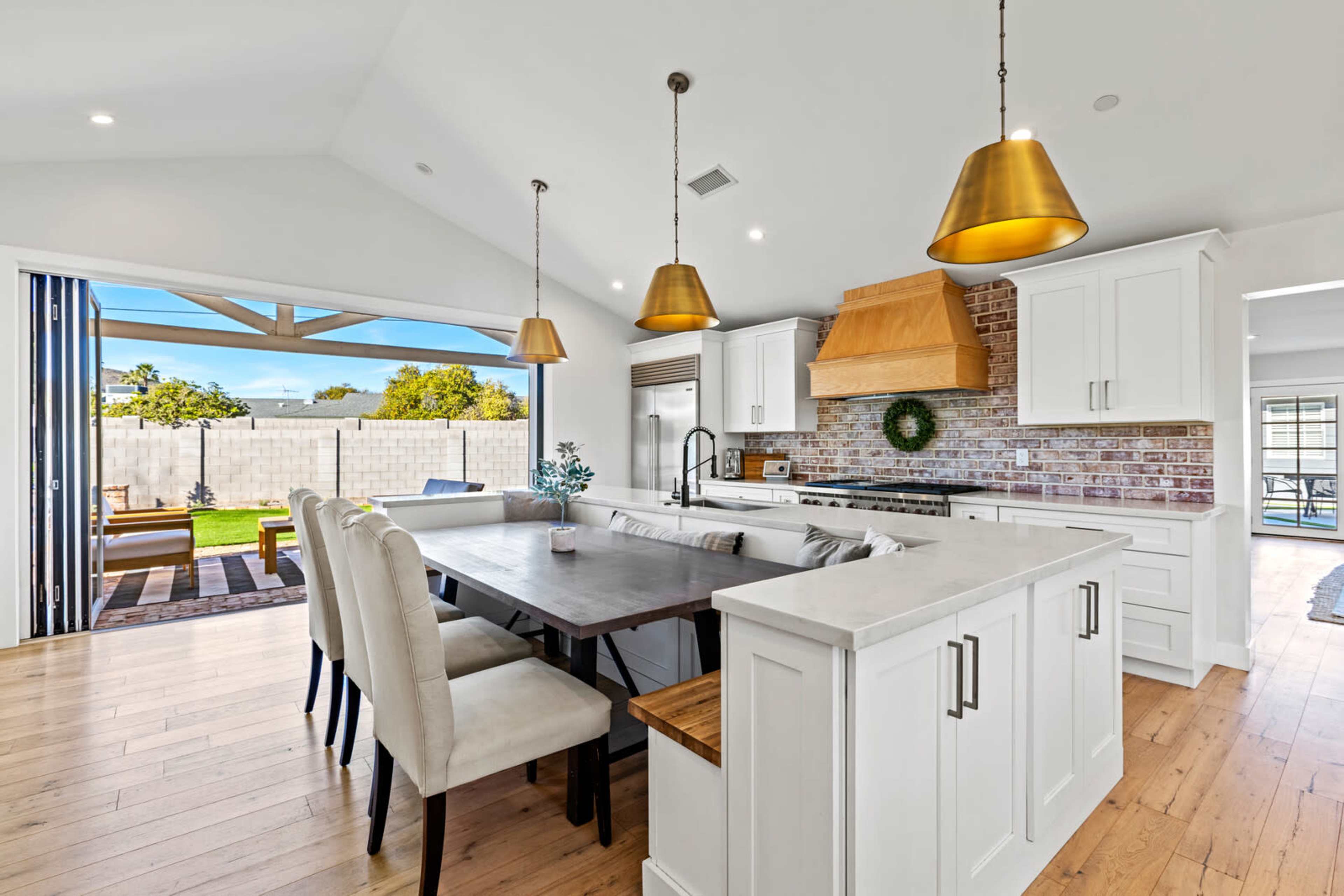 A modern kitchen features a large dining table, white cabinetry, wooden accents, and pendant lights, with a view of a backyard through sliding glass doors.