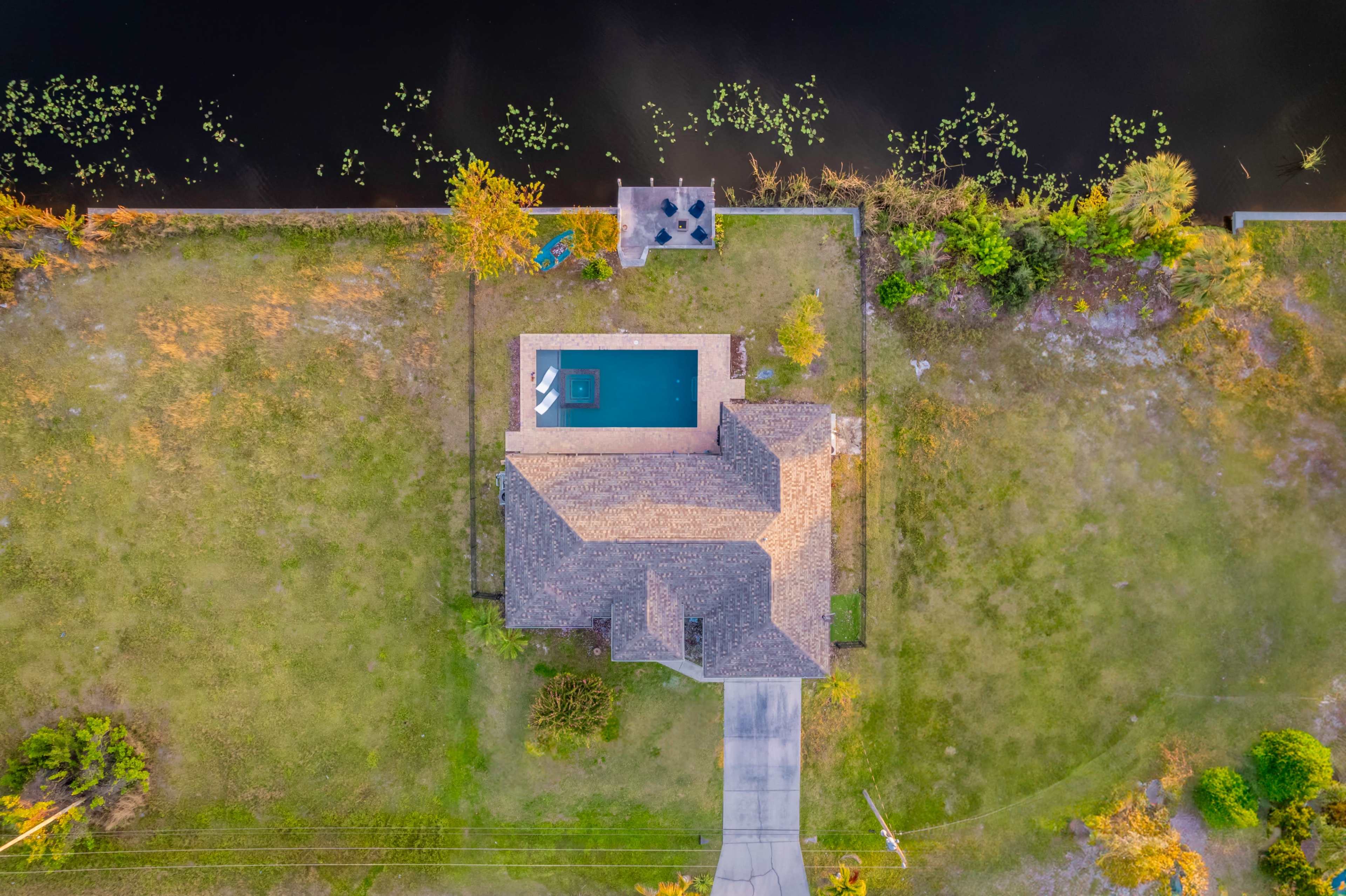 An aerial view of a single-story house with a swimming pool, situated beside a dark water body, surrounded by a grassy yard.