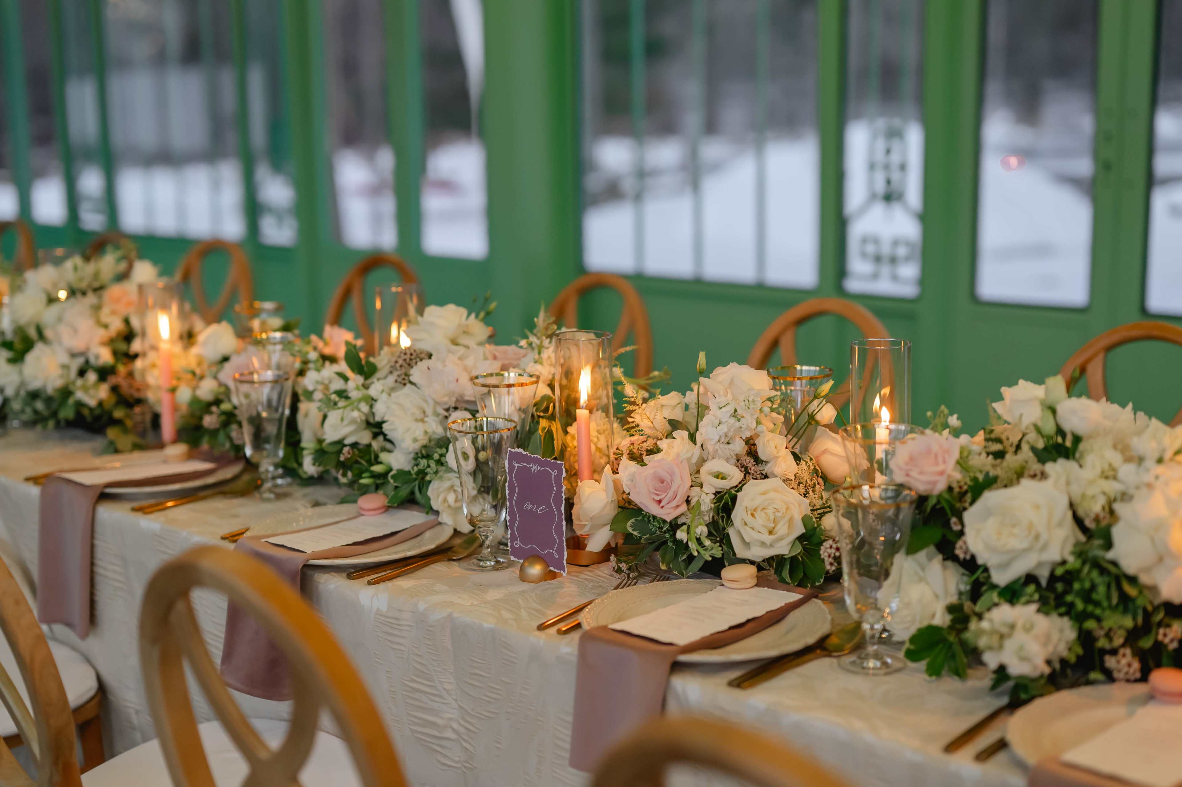 A long dining table is elegantly set with floral centerpieces, candles, and place settings in a room with green windows.