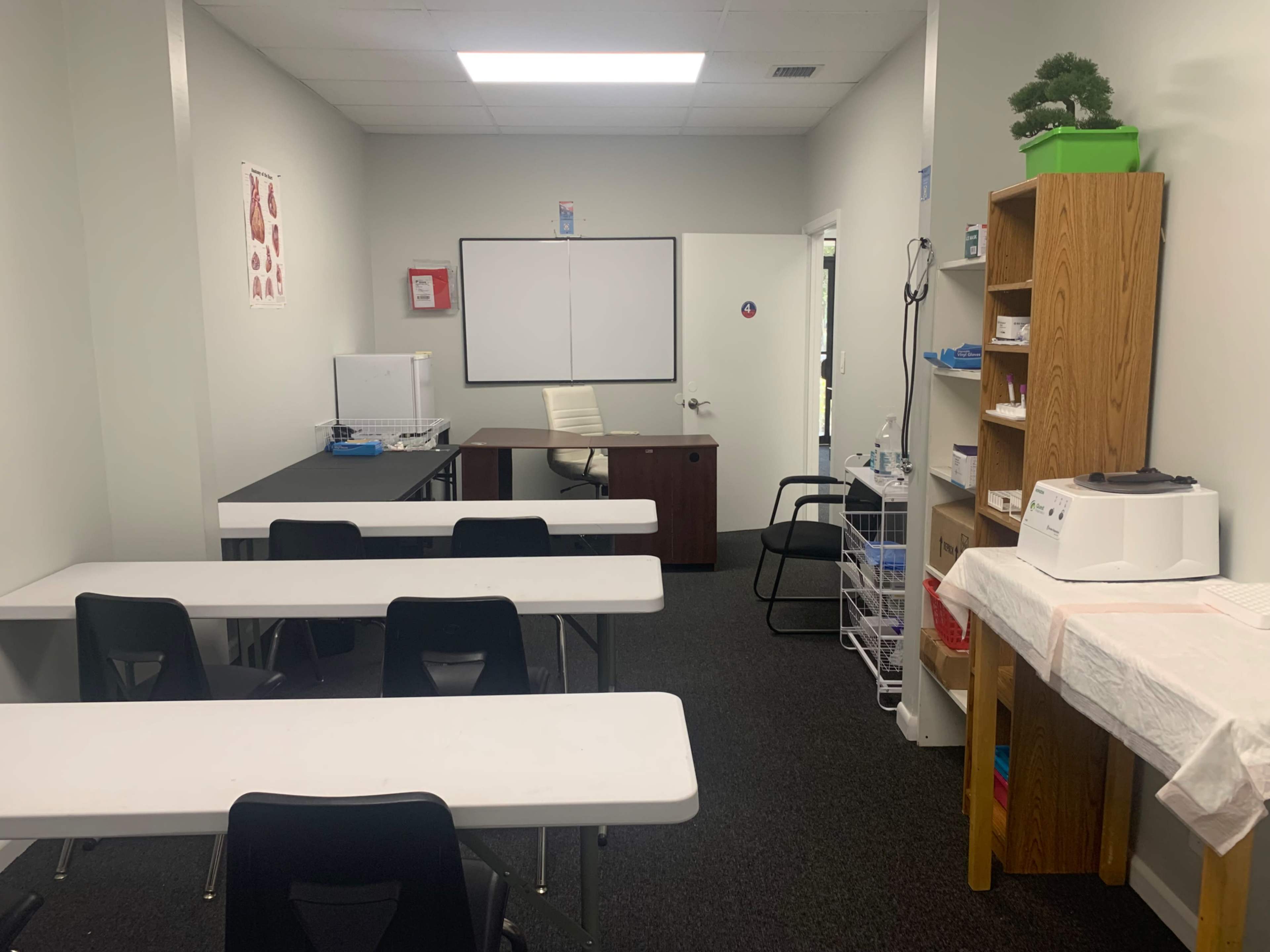 A classroom with several white tables and black chairs, a desk in the corner, and shelves containing various items.