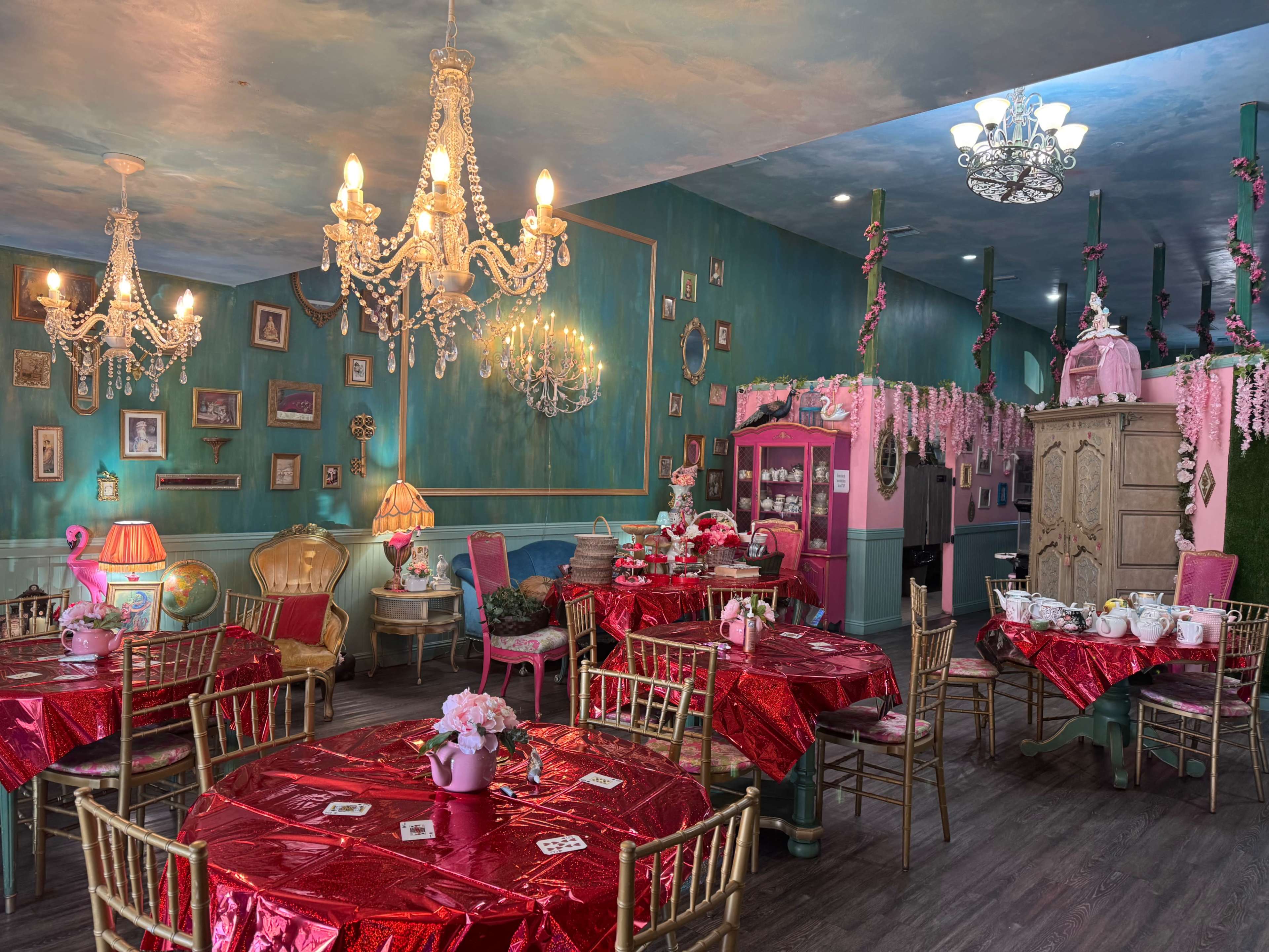 The image depicts a vibrant tea room with pink walls, ornate chandeliers, and red tablecloths decorated with floral arrangements.