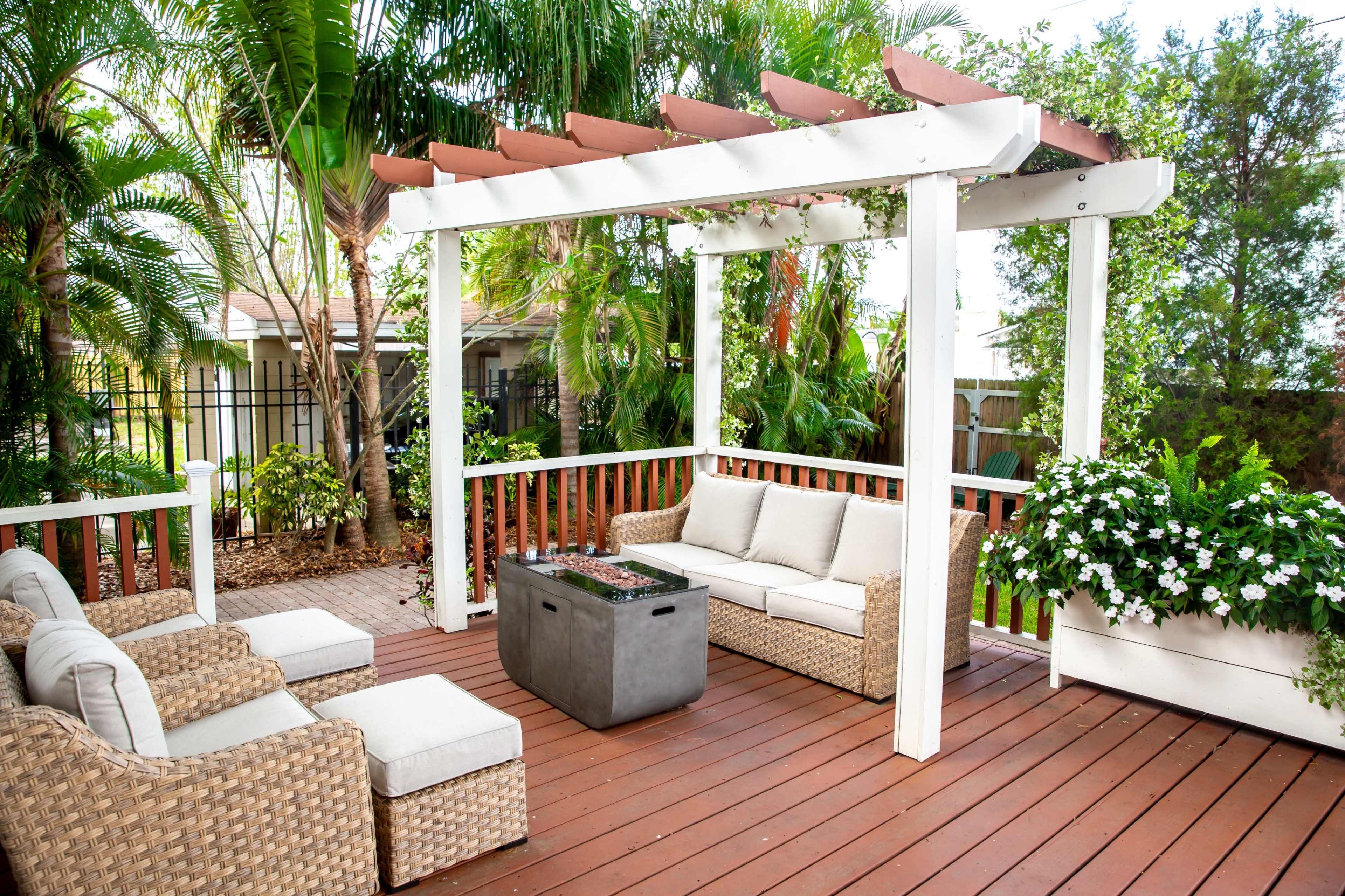 The image shows a landscaped outdoor patio featuring a seating area with wicker furniture, a central fire pit, and surrounding greenery.