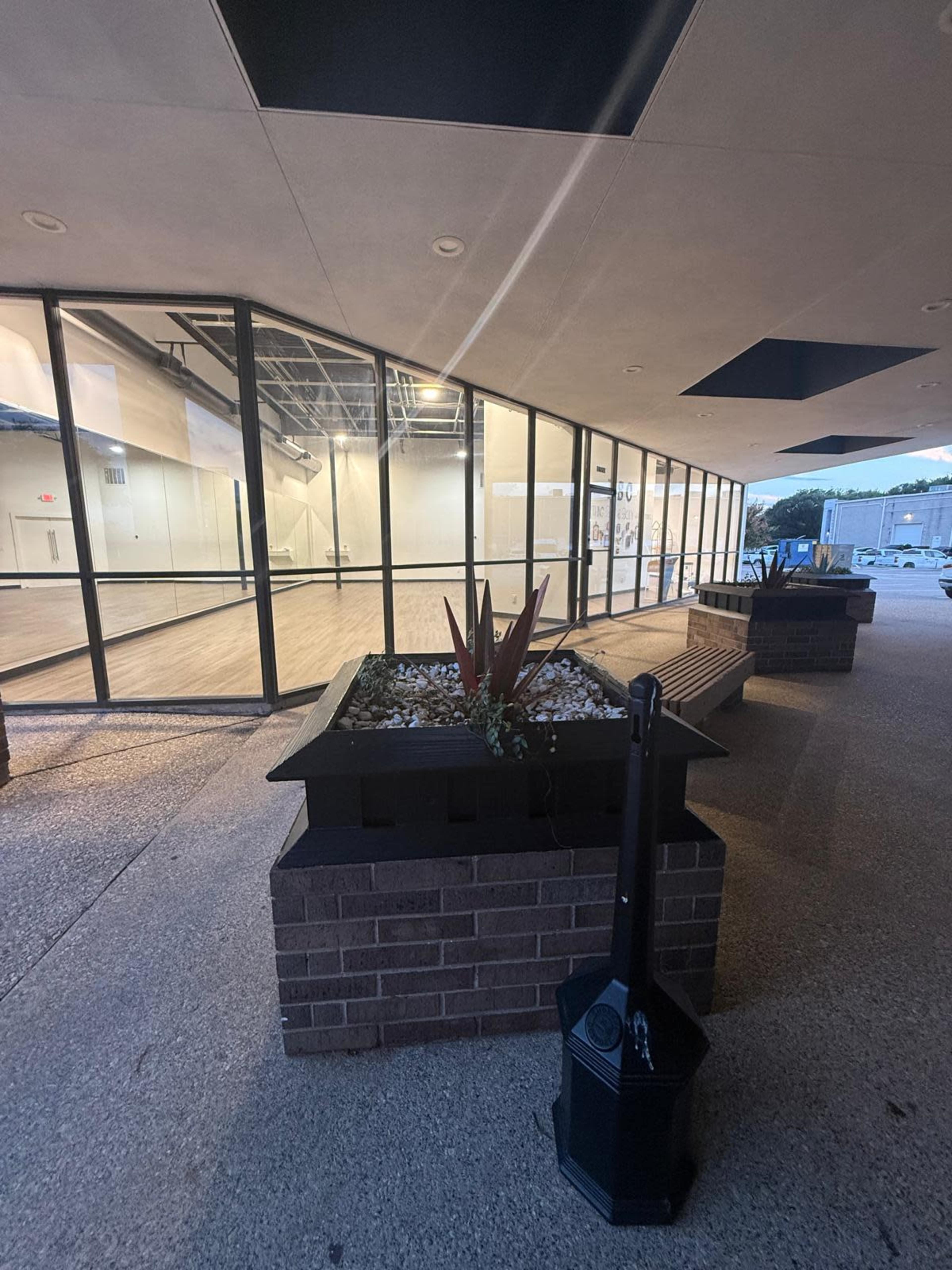 The image shows a modern building with large glass windows, a stone planters filled with plants, and a sidewalk leading to an indoor space.