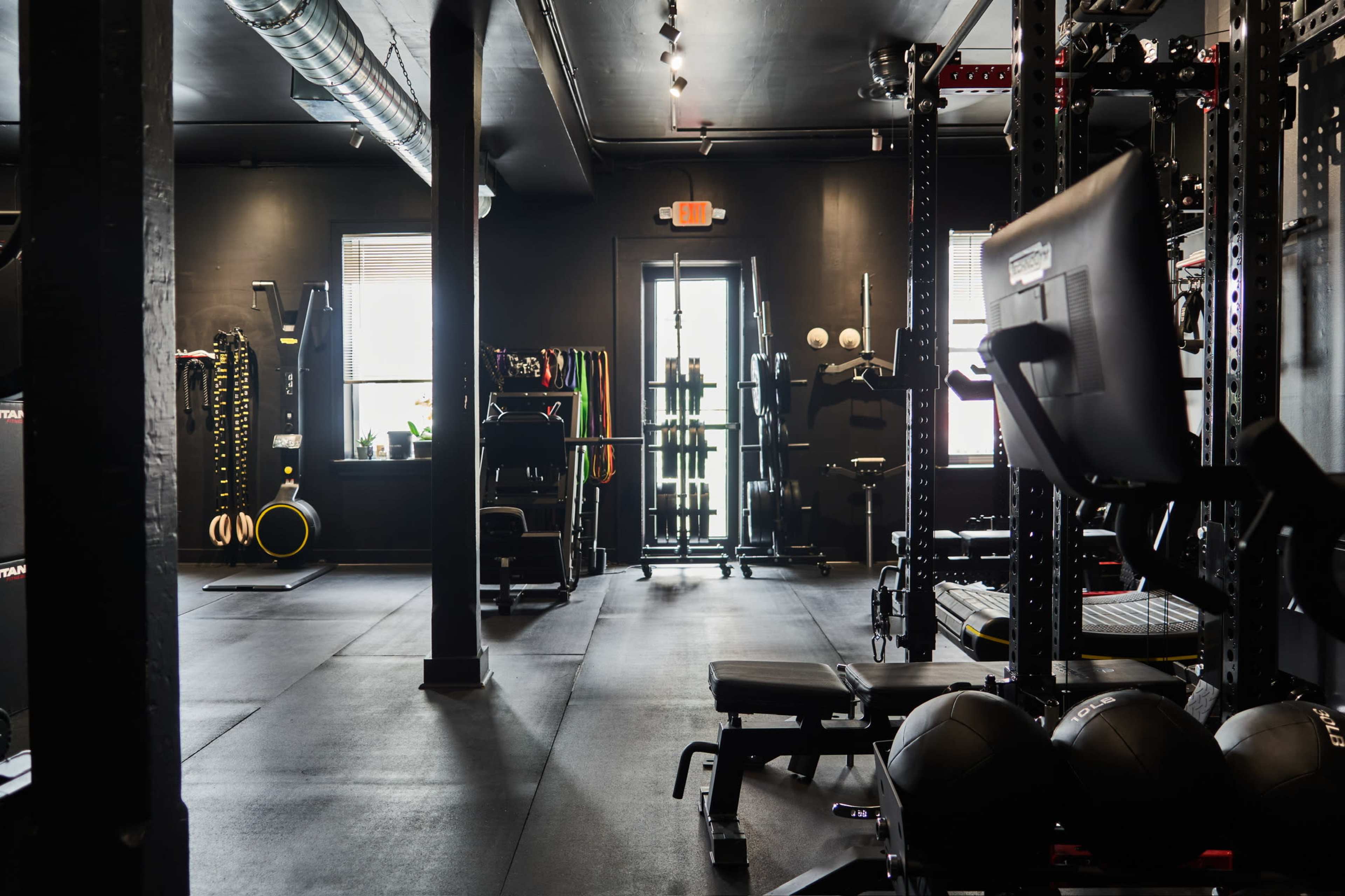 The image shows a dimly lit gym with various workout equipment, including weightlifting benches, resistance bands, and exercise balls, arranged neatly in an open space.