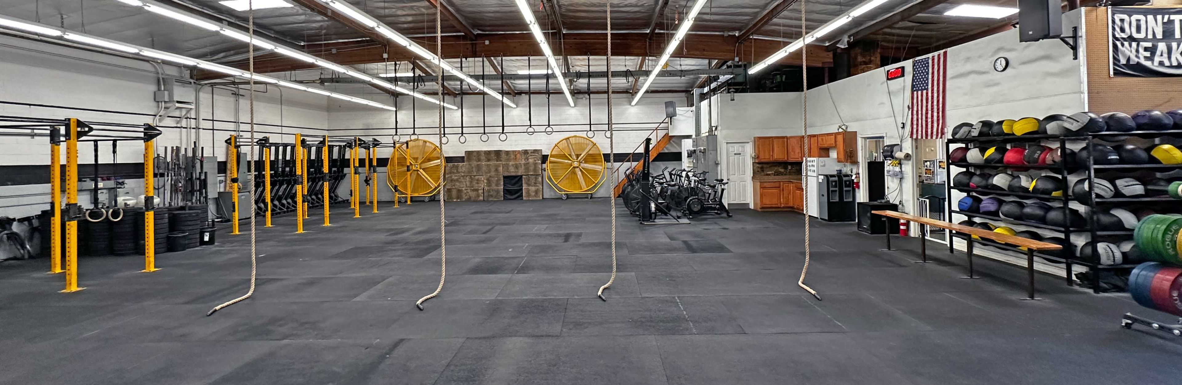 The image depicts a large, open gym space with various exercise equipment, including weights, bikes, and gymnastic rings hanging from the ceiling.