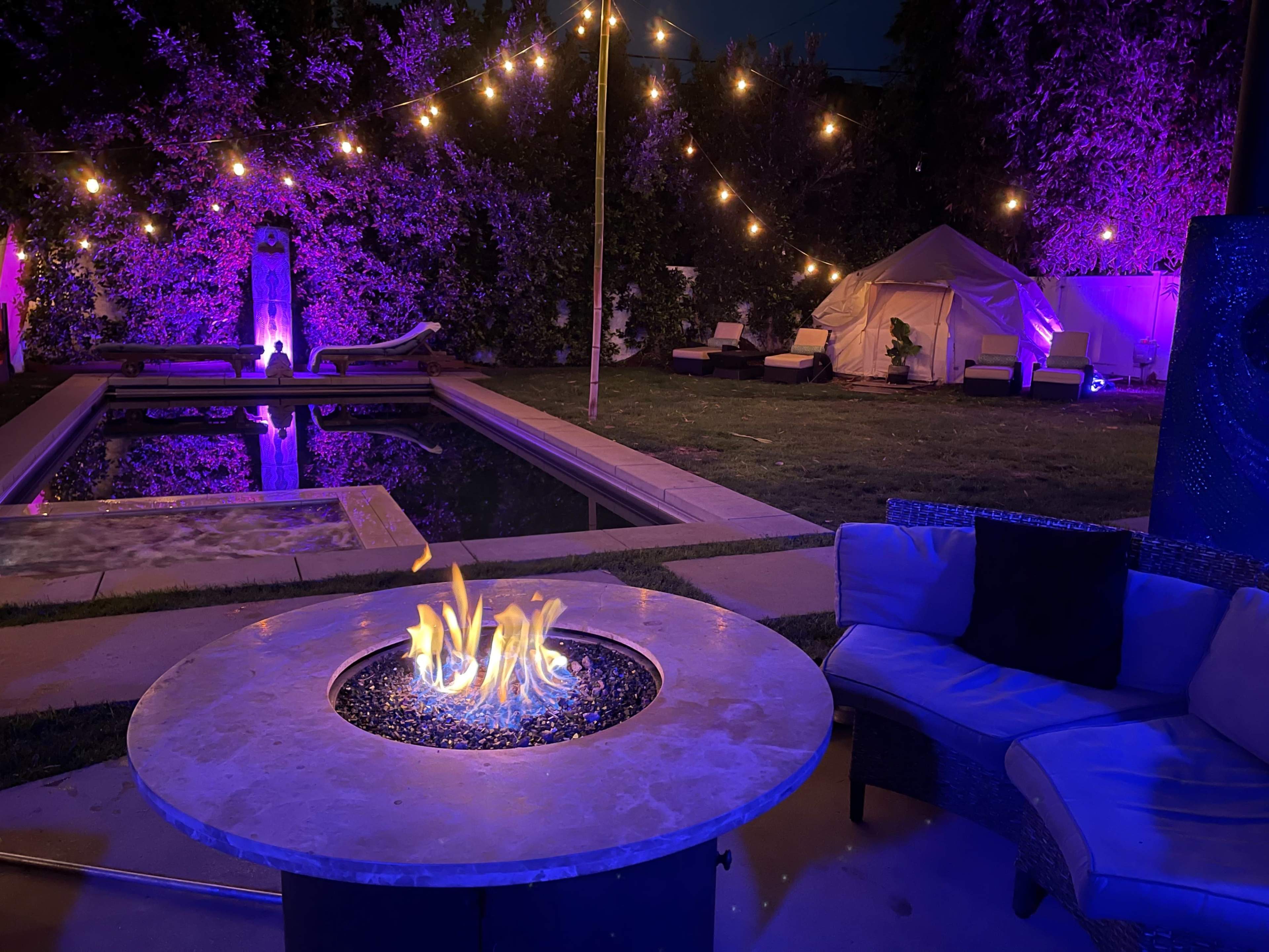 A fire pit with flames sits in the foreground, surrounded by seating, while a pool and illuminated tent are visible in the background under string lights.