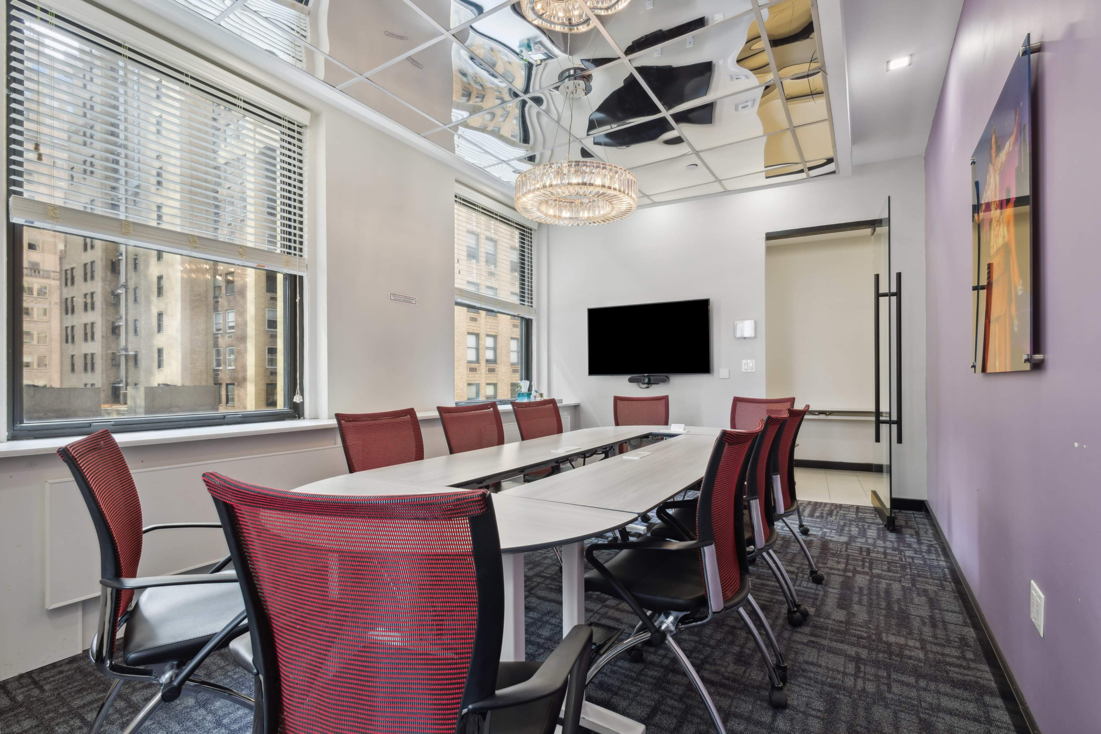 A conference room features a long table surrounded by red chairs, with large windows allowing natural light and a mirrored ceiling.