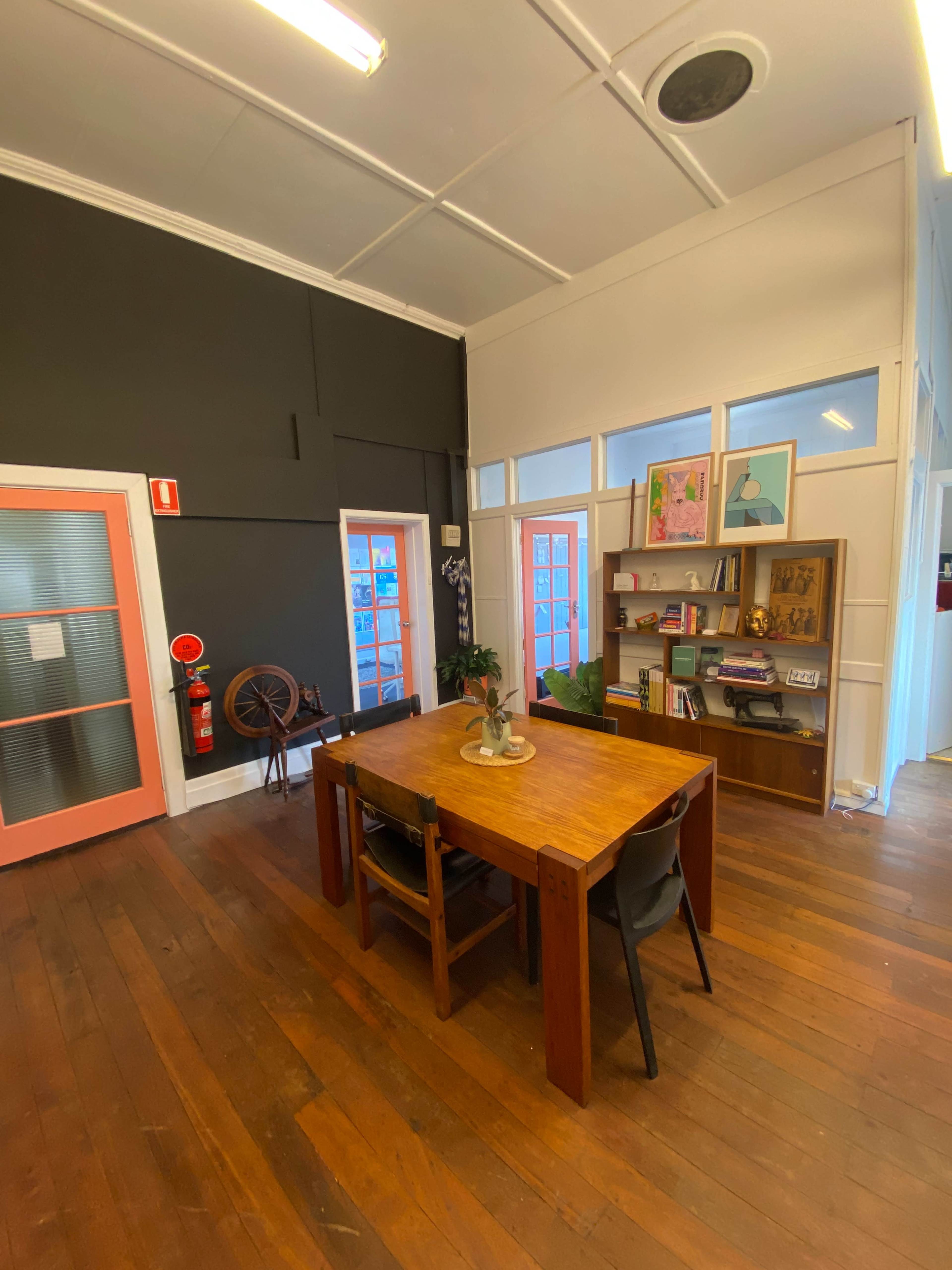 The image shows a bright, modern office space featuring a wooden table surrounded by chairs, a bookshelf filled with books and decor, and walls painted in contrasting colors.
