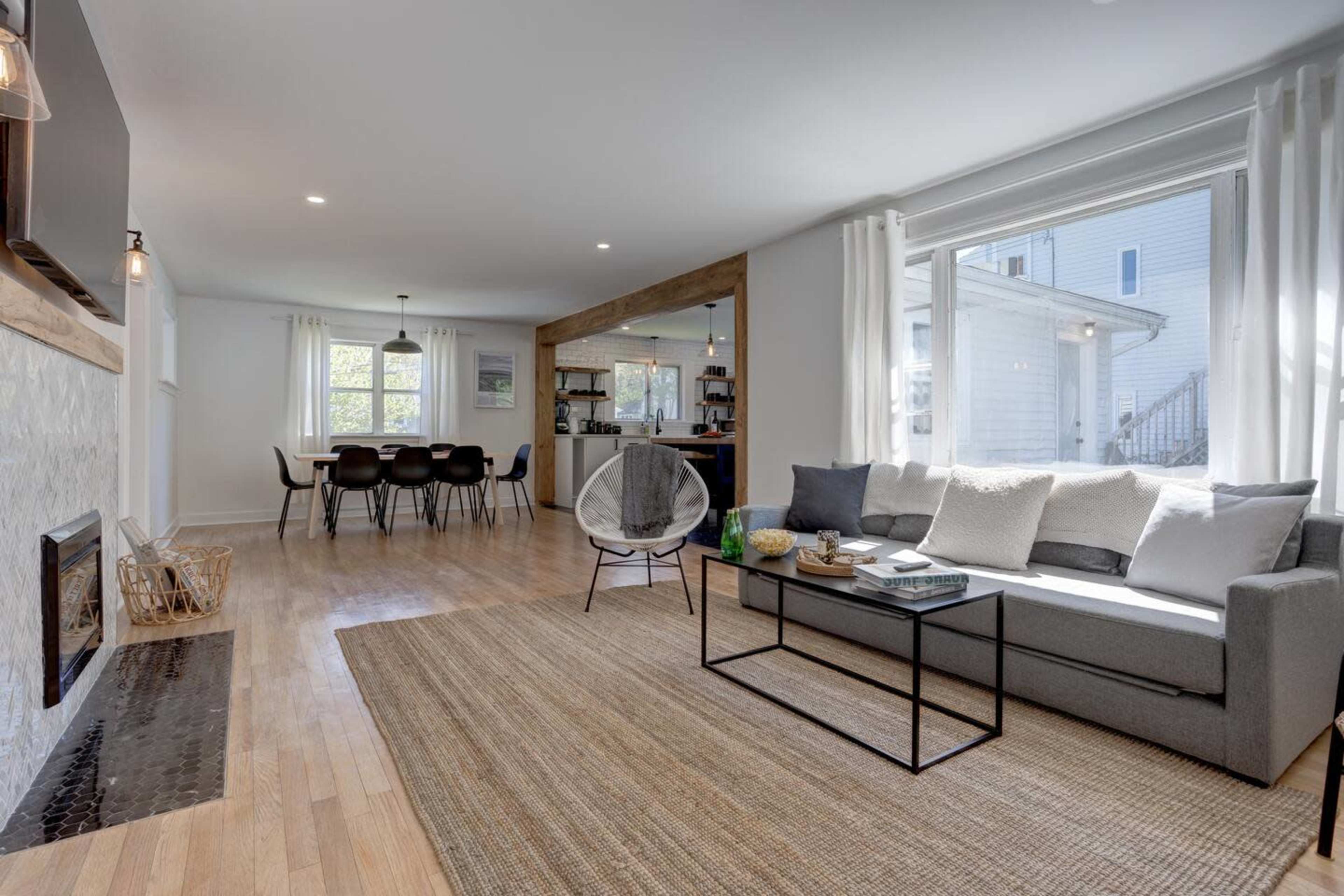 A bright living room featuring a gray sofa, a black coffee table, a large window, and a view into a dining area with chairs.