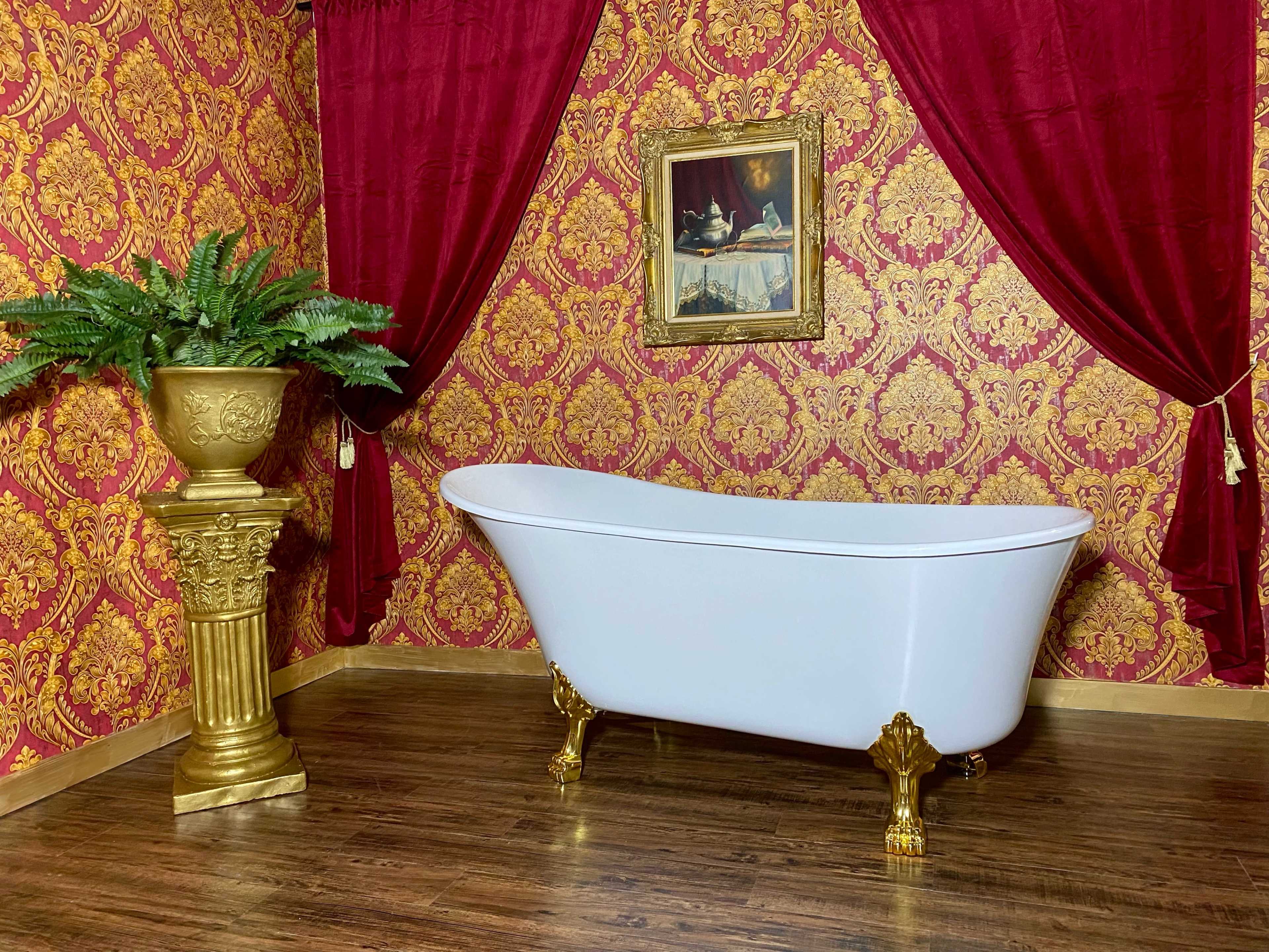 A vintage clawfoot bathtub is positioned against a richly patterned red and gold wallpaper, accompanied by a decorative gold plant stand and a framed painting.