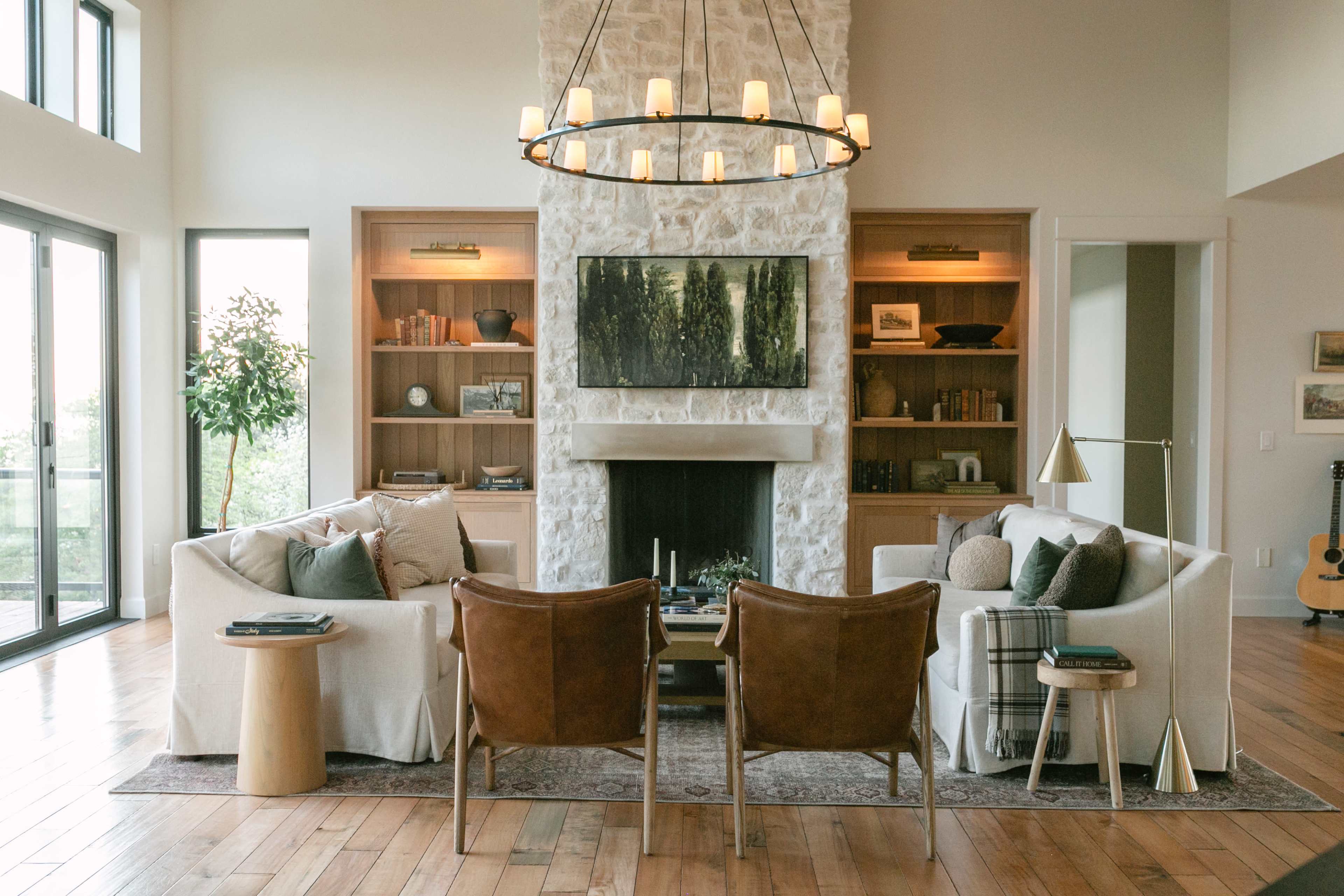 The image shows a spacious living room featuring two armchairs, a stone fireplace, built-in bookshelves, and large windows allowing natural light to enter.