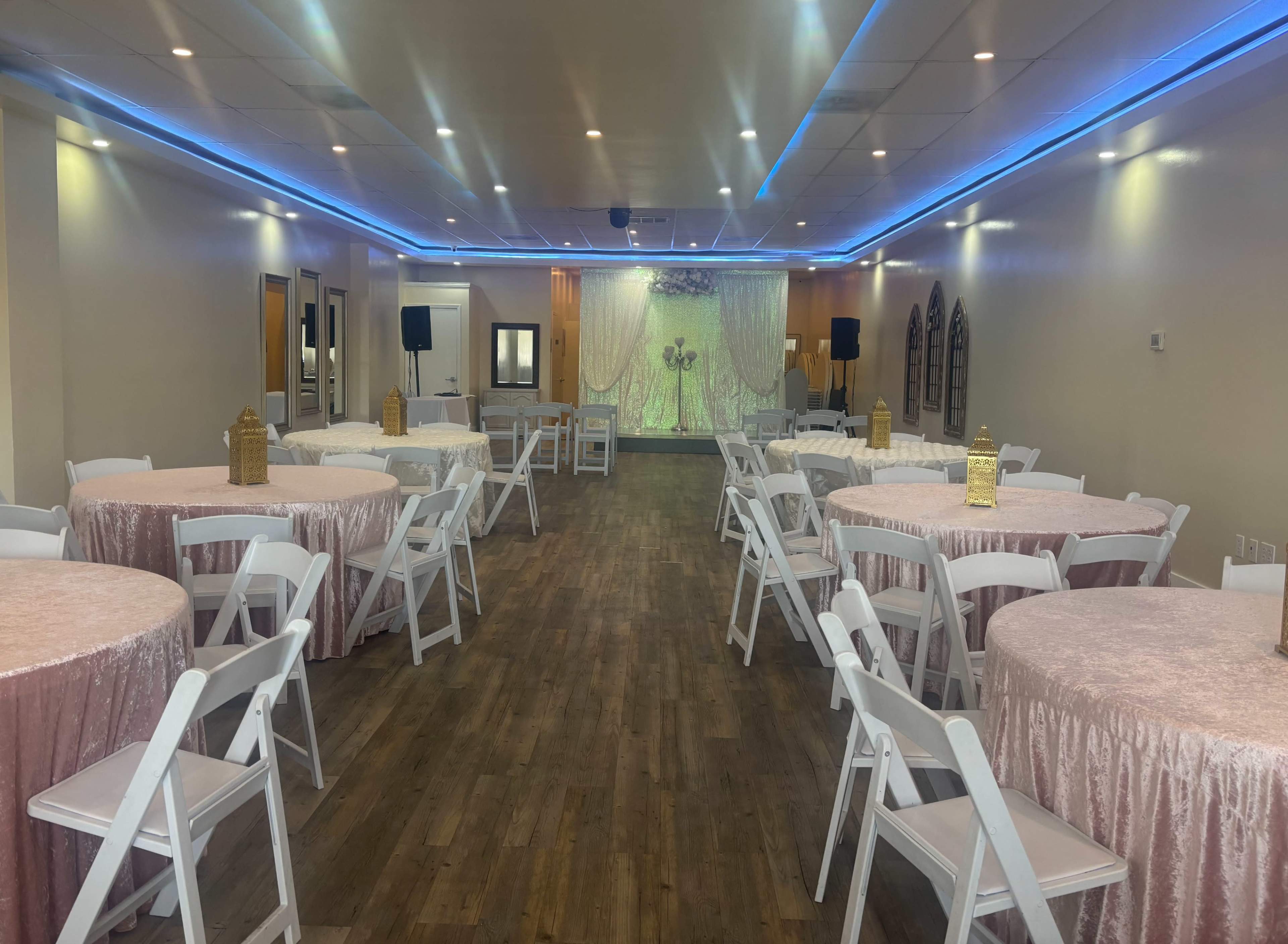 The image shows a spacious event venue arranged with round tables covered in pink tablecloths and white chairs, illuminated by blue LED lighting.