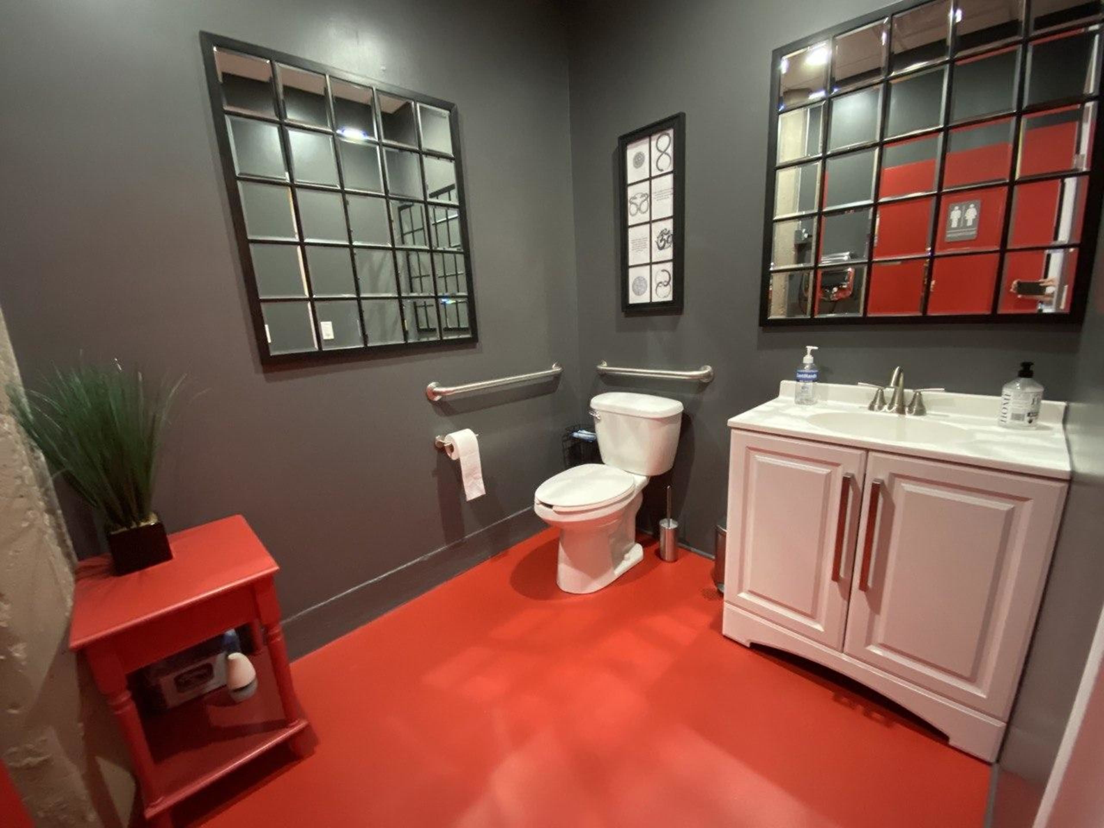 A small bathroom with a red floor, a white vanity, and a toilet, accompanied by two large mirrors and a decorative wall frame.