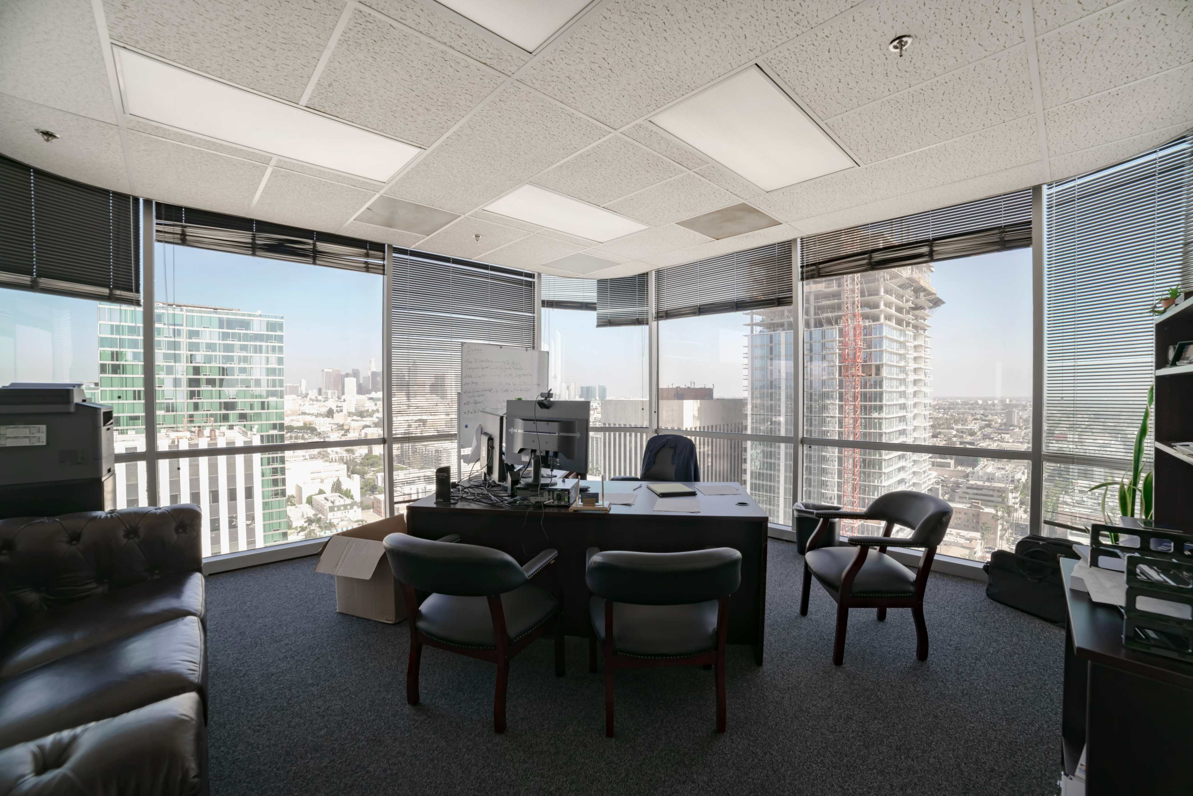 A modern office space with large windows offering a view of a city skyline, equipped with a desk, chairs, and a computer setup.