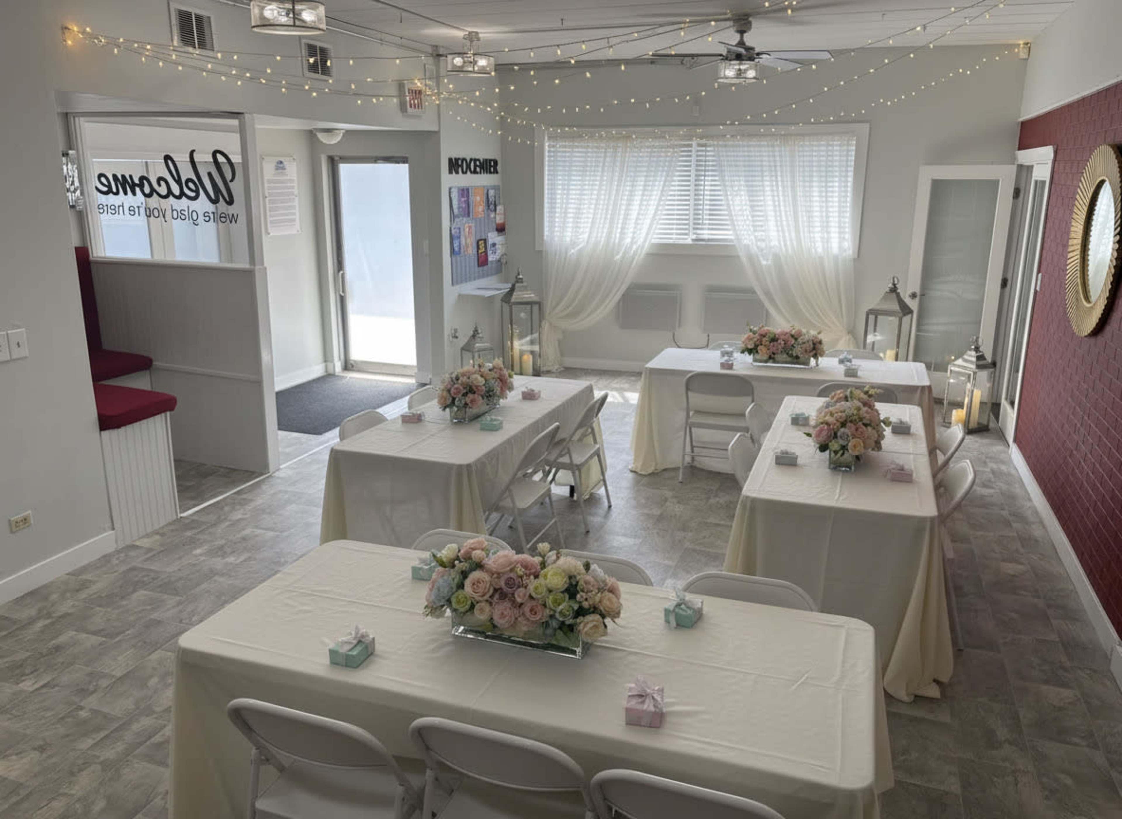 The image shows a decorated room set up for an event, featuring several tables with floral centerpieces and festive lighting.