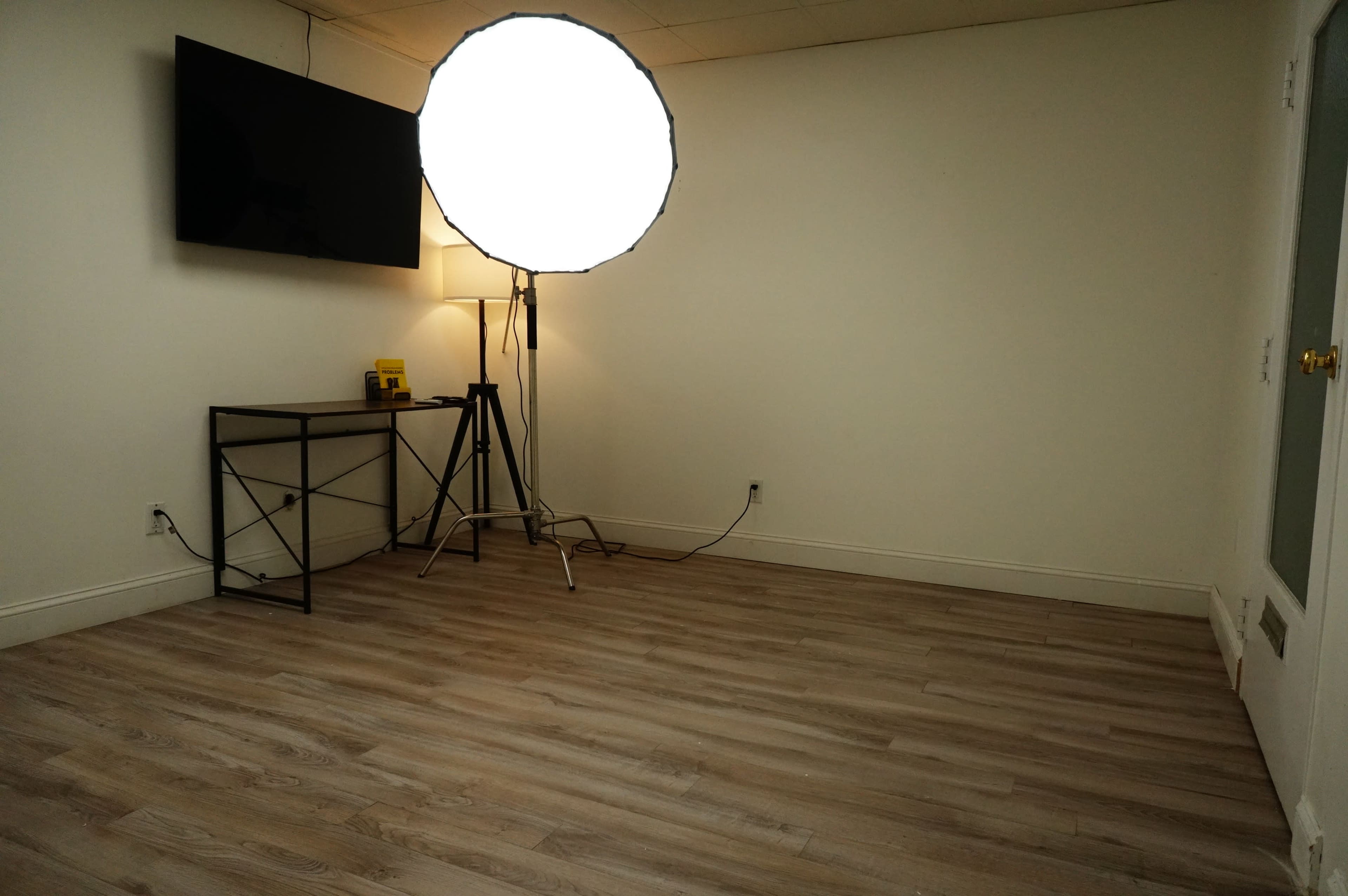 A sparsely furnished room with light wooden flooring, a large round softbox light, a standing lamp, a small table, and a wall-mounted TV.